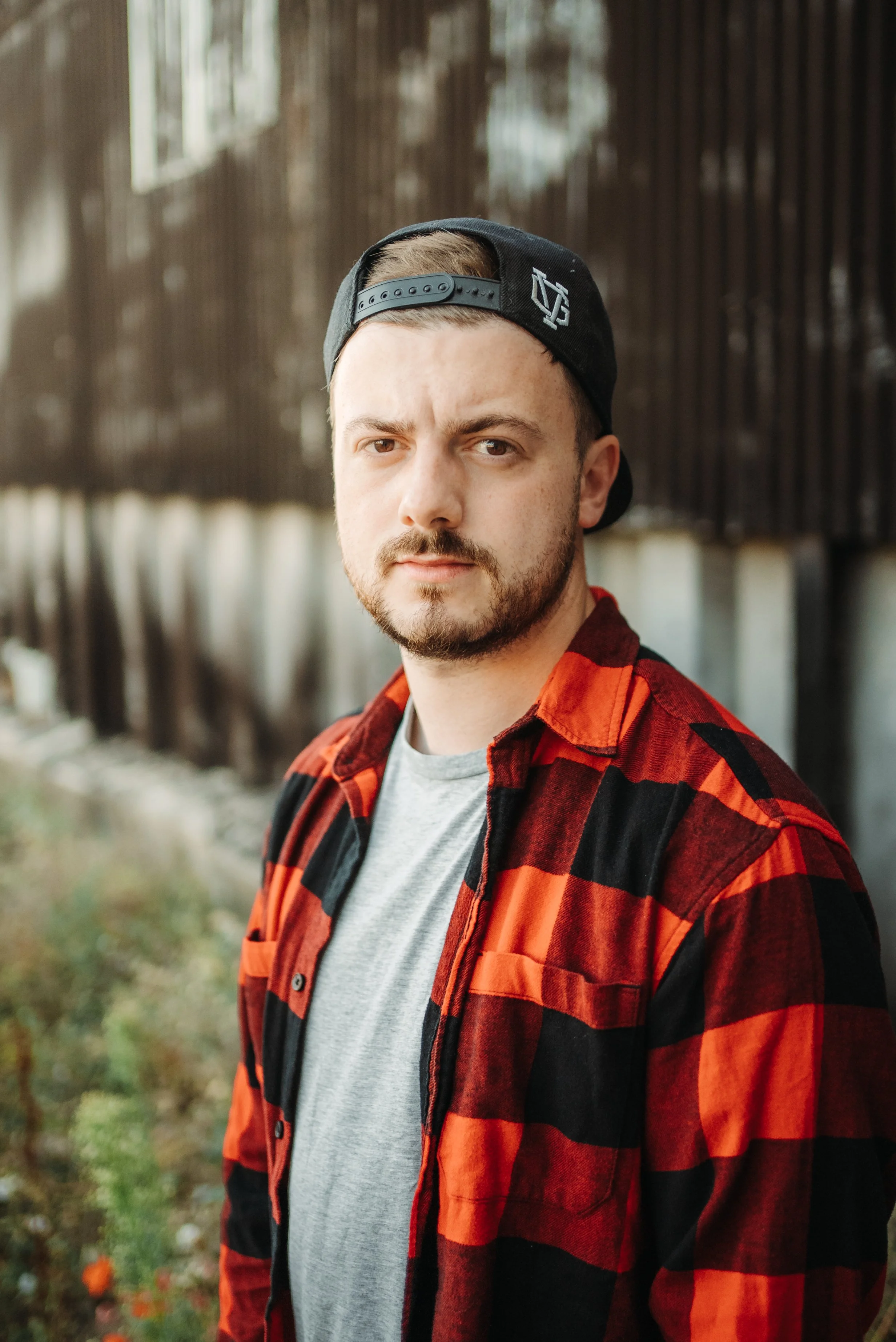 A young man with a beard and mustache wearing a backward black baseball cap, gray t-shirt, and red and black plaid shirt, standing outdoors near a fence.