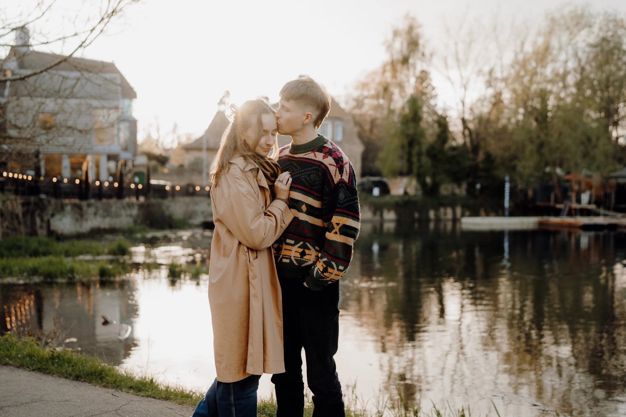 A couple stands close by a river, with the man kissing the woman's forehead as she gently touches his chest, during sunset with trees and houses in the background.