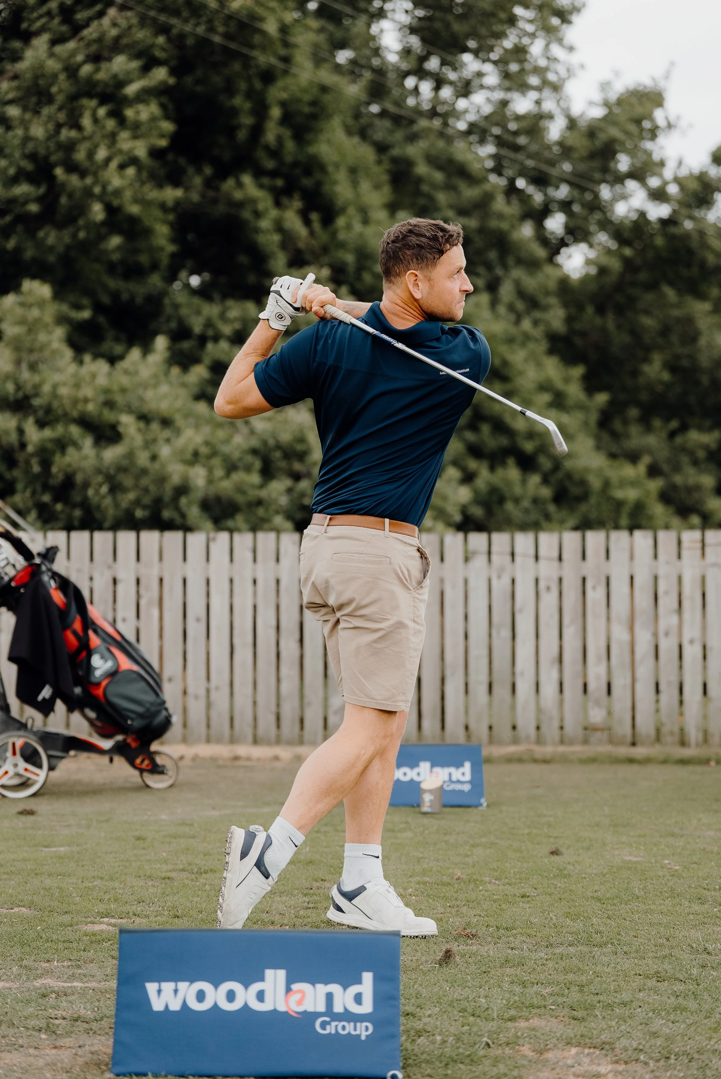 Man wearing navy polo shirt and beige shorts swinging golf club on a golf course with wooded background, golf bag, and 'woodland Group' sign in the foreground.