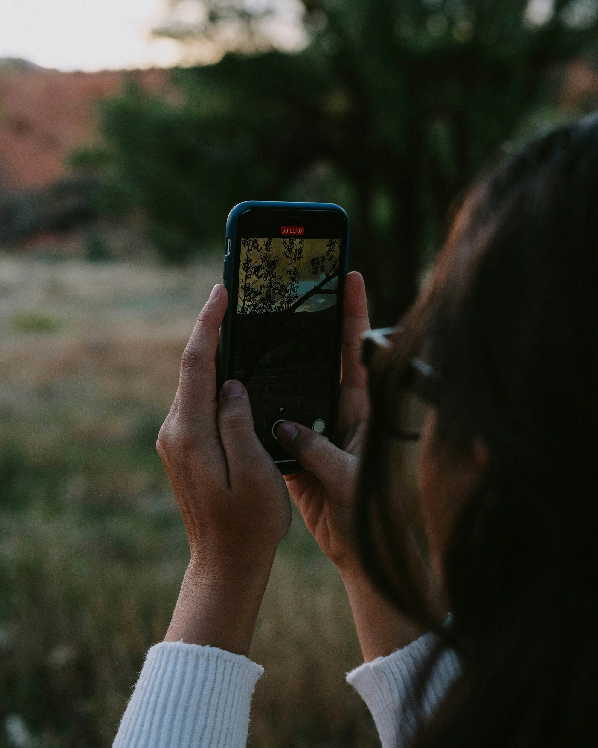 A person holding a smartphone, recording or taking a picture of a landscape with trees.