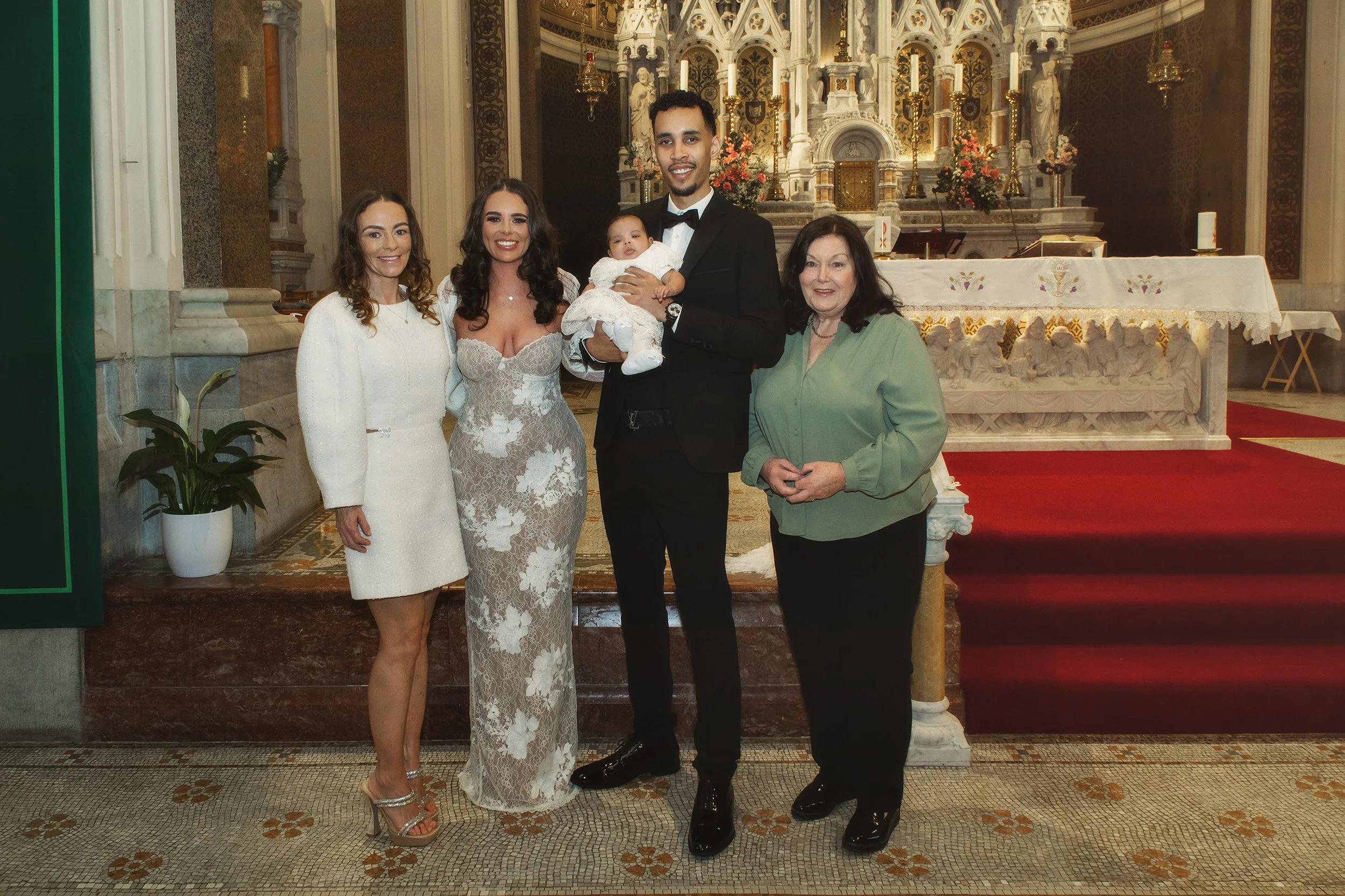 A group of five people, including a woman in a white dress holding a baby, a man in a black tuxedo, and three women, are posing inside a church with ornate altar and altar table in the background.