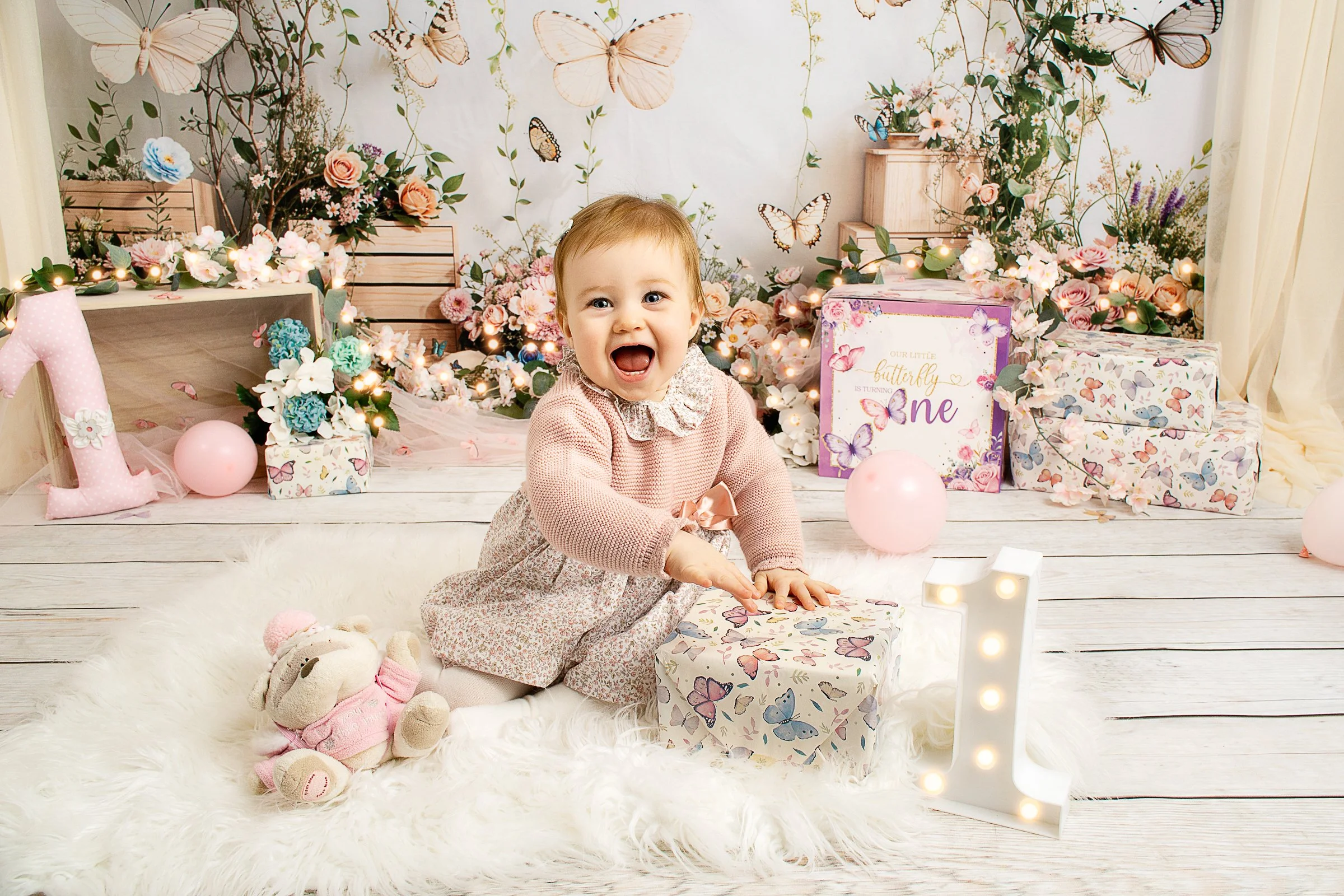 A baby girl with short light brown hair wearing a pink cardigan and floral dress, sitting on a white fluffy rug, smiling with her mouth open, surrounded by pink and white balloons, plush toys, and floral wrapped gifts for a butterfly-themed celebration in a decorated room with butterfly wall art and floral arrangements.