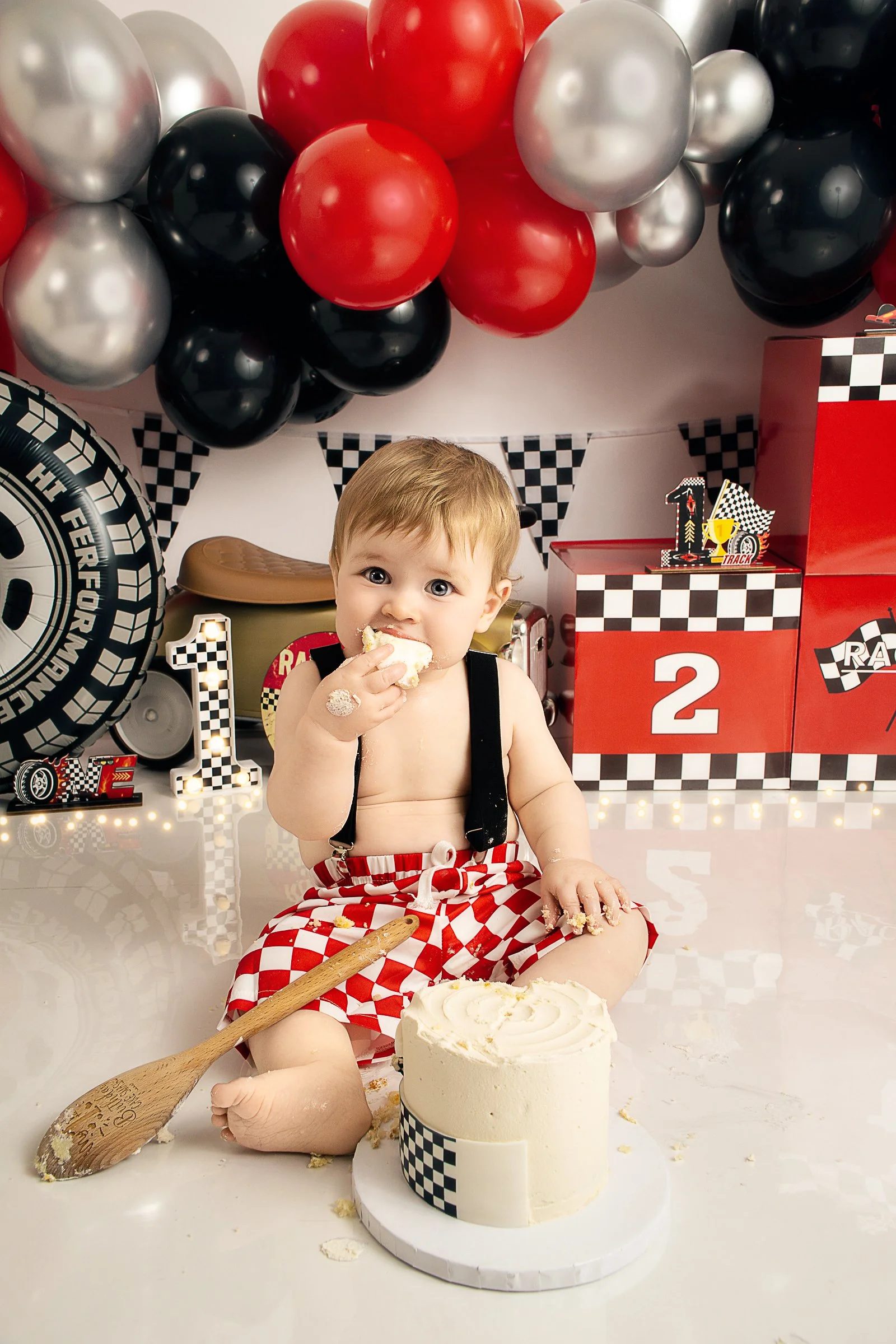 A baby boy wearing red and white checkered shorts, sitting in front of a white birthday cake with white frosting. He is eating cake with his hand, with cake on his face and hands. The background features red, black, silver balloons, and a racing-themed decorations with checkered flags and racing cars.