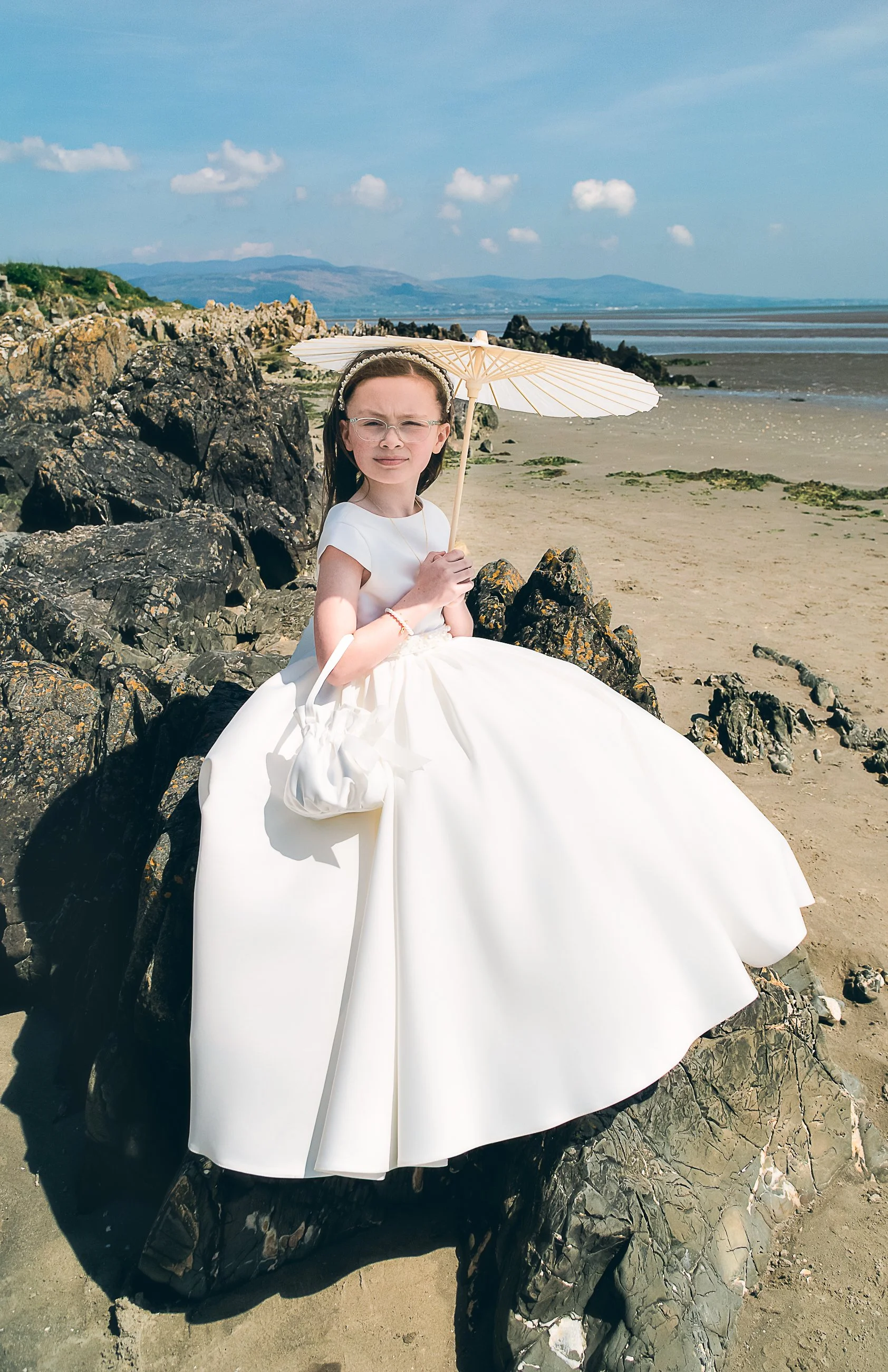 A young girl in a white dress holding a white parasol, sitting on rocks at the beach with sand, rocks, and ocean in the background.