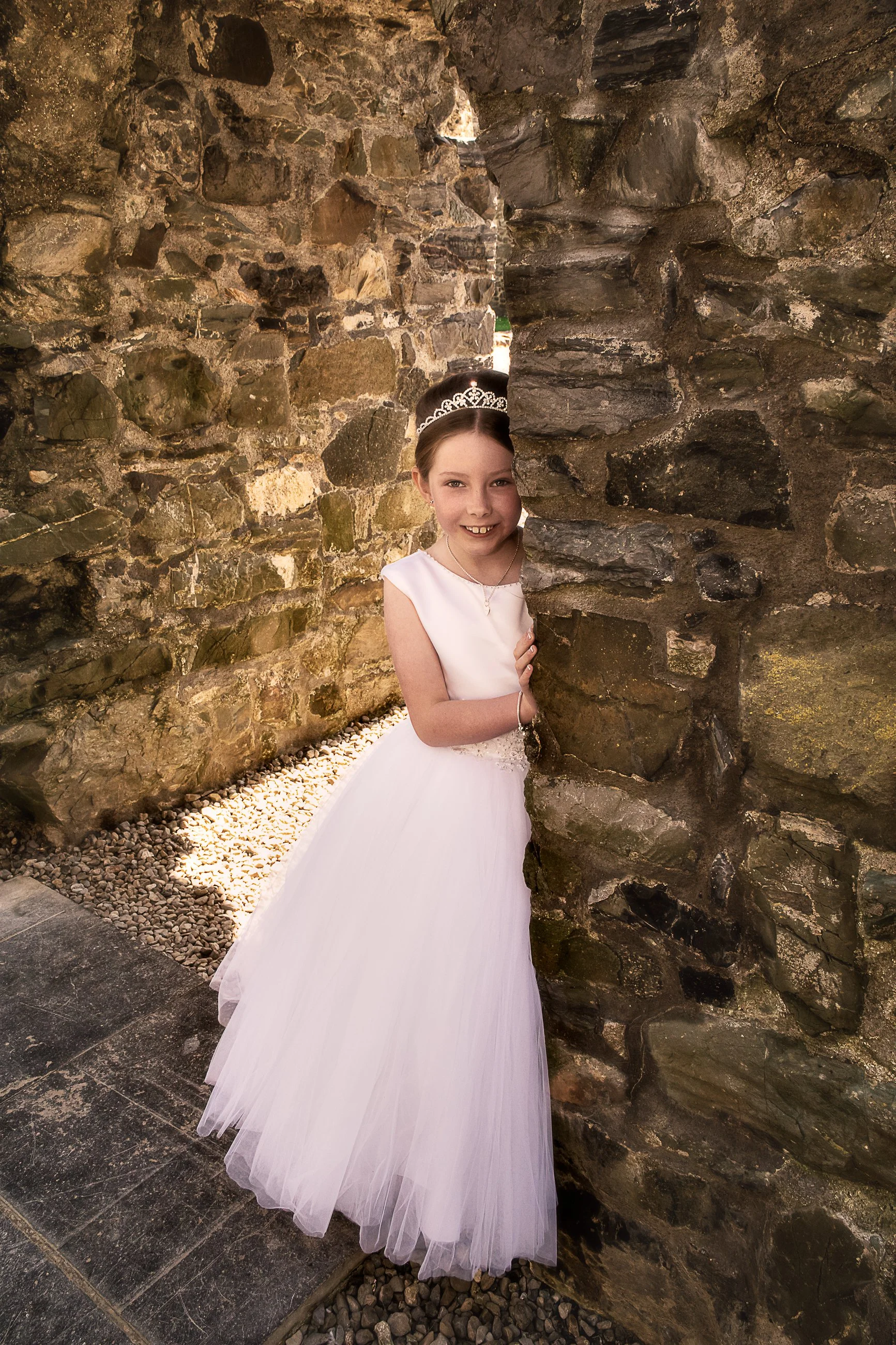 A young girl in a white dress and tiara posing between two stone walls.