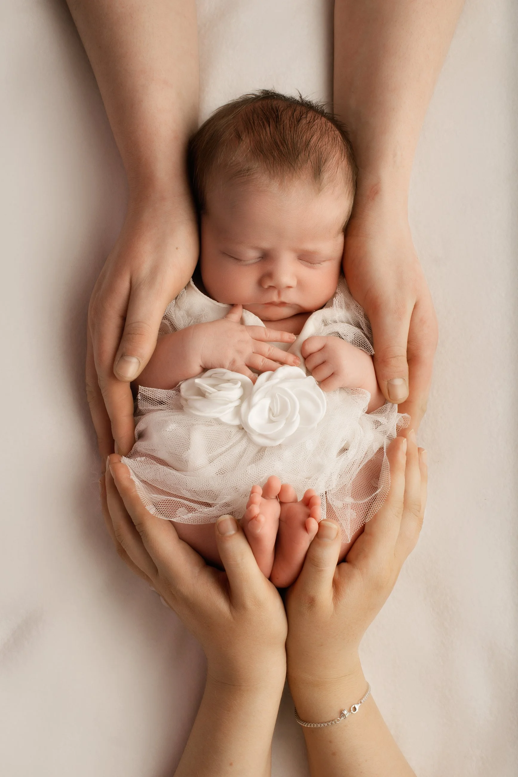 A newborn baby sleeping peacefully in the hands of a person, wearing a white dress with floral accents, surrounded by the person's hands and arms.