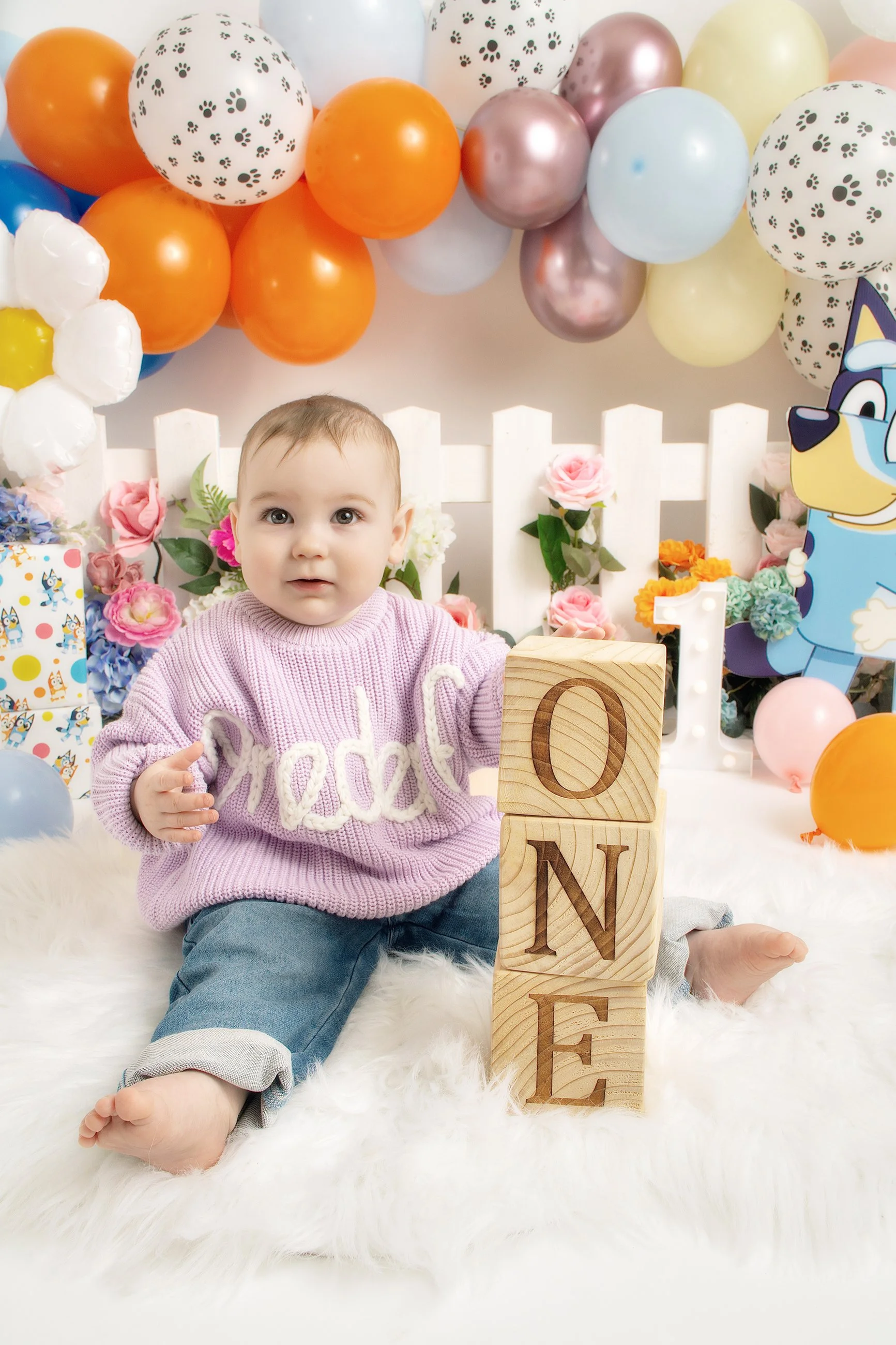 A young child sitting on a white furry rug, surrounded by colorful balloons and flowers, holding wooden blocks spelling 'ONE', celebrating a first birthday.