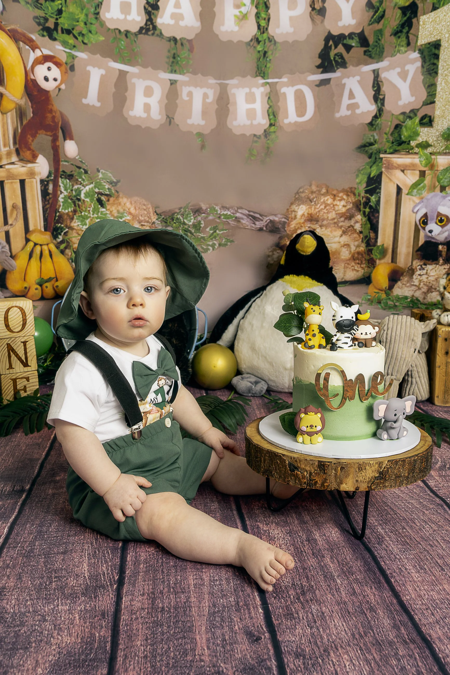 A young child with blue eyes, wearing a green hat, white t-shirt with a monkey graphic, green overalls, and a bowtie, sitting on a wooden floor in front of a birthday cake decorated with animal toppers, with a jungle-themed backdrop and birthday decorations.