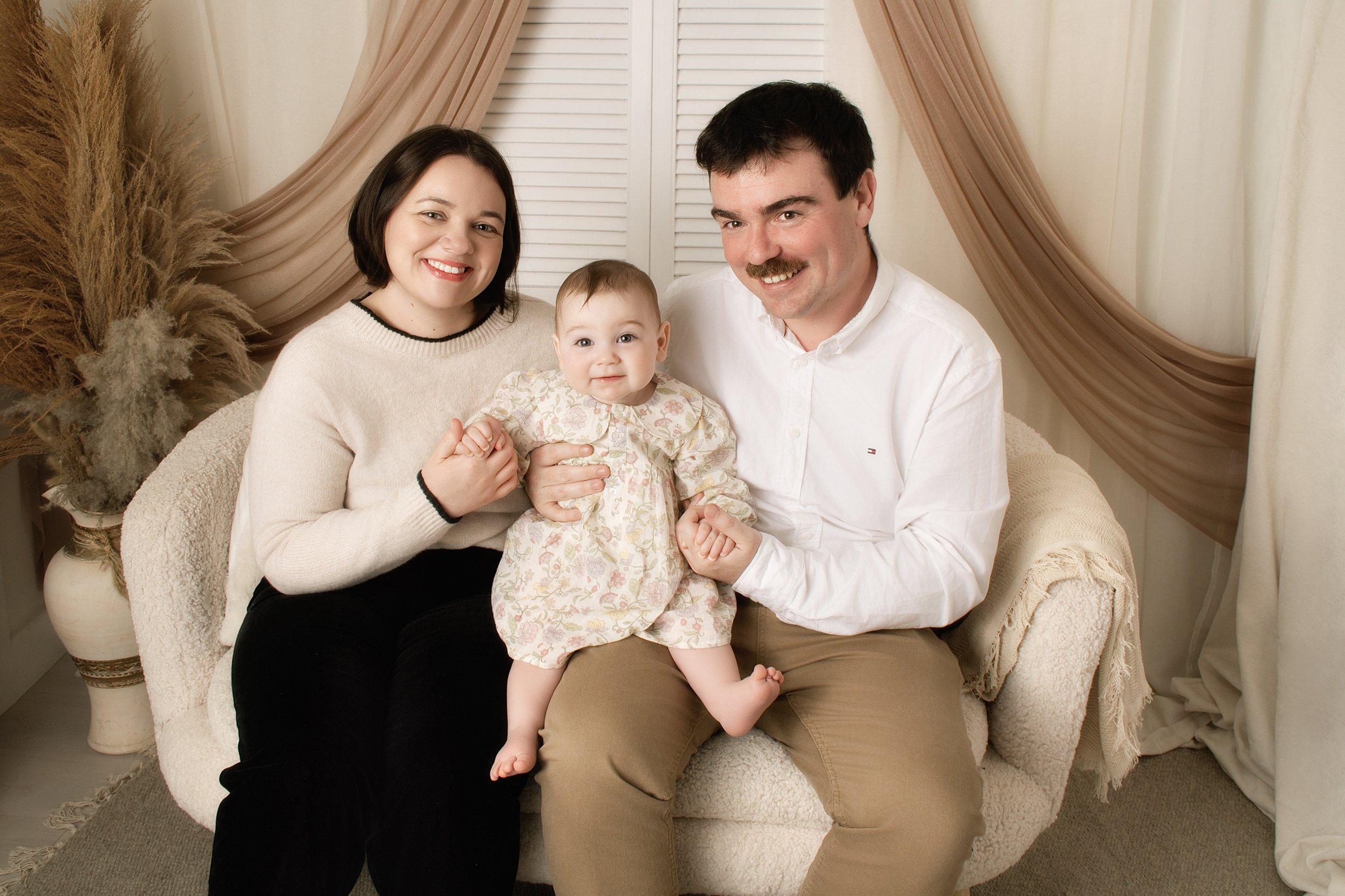 A family of three, including a woman, a man, and a baby girl, sitting on a beige couch in a cozy living room with beige curtains and pampas grass in a vase nearby.