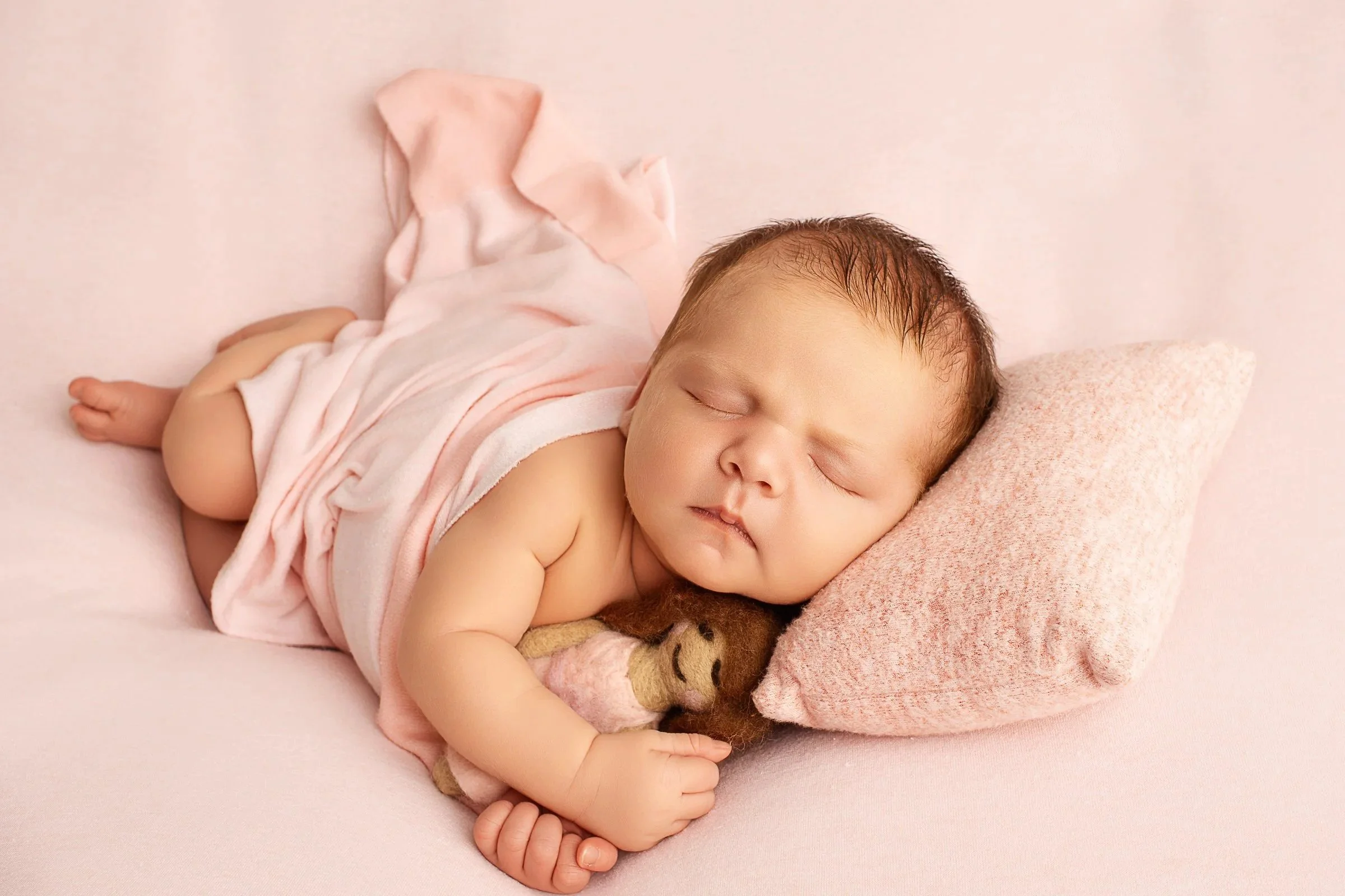 A sleeping baby with light brown hair, lying on a pink bed, hugging a small stuffed tiger toy.