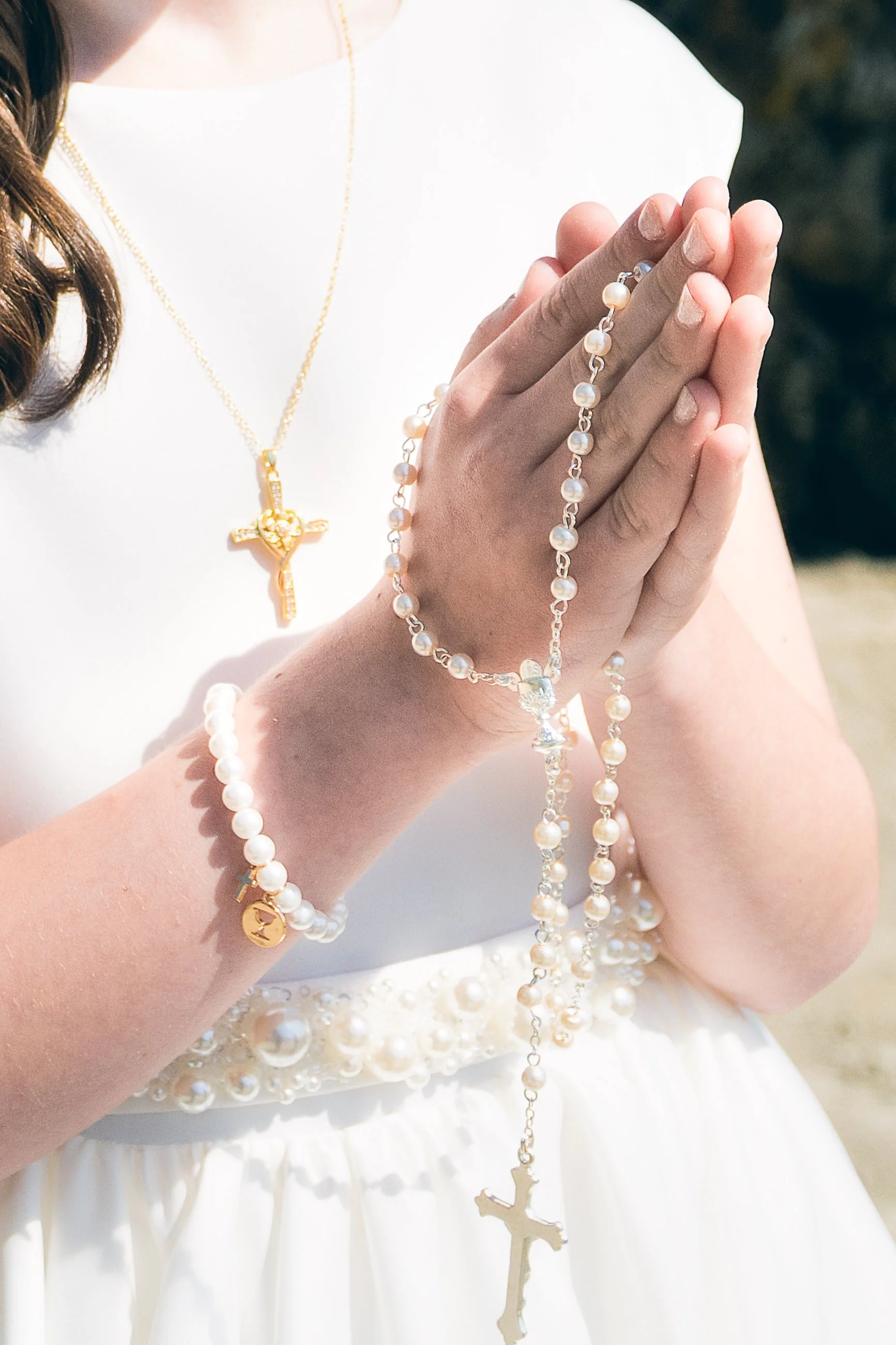 A bride holding her hands together with pearl jewelry and a gold cross necklace, wearing a white dress with pearl embellishments.