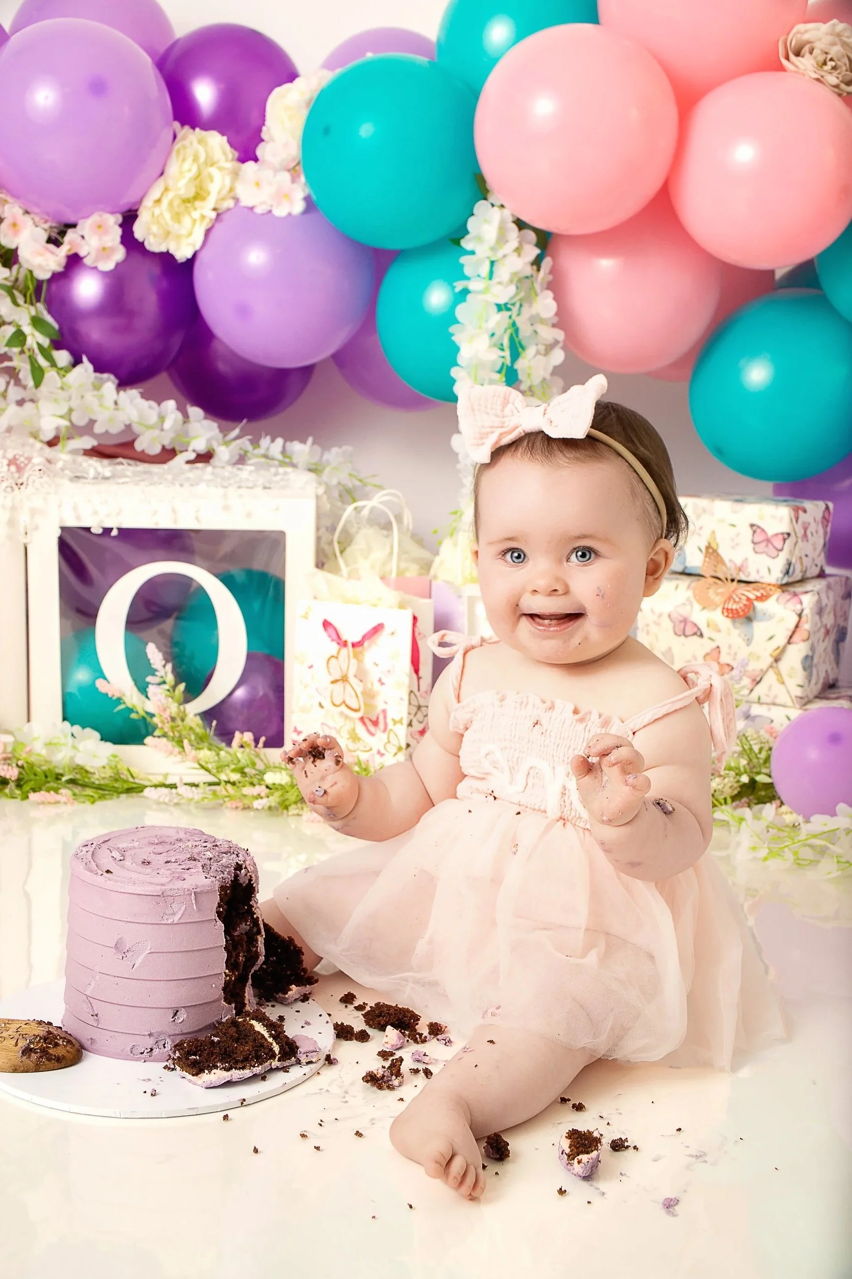 A young girl with blue eyes, wearing a pink dress and a headband with a bow, sitting at a birthday party with purple, pink, and teal balloons in the background. She has a cake smash with chocolate cake and purple frosting on a white table, surrounded by scattered cake pieces and crumbs.