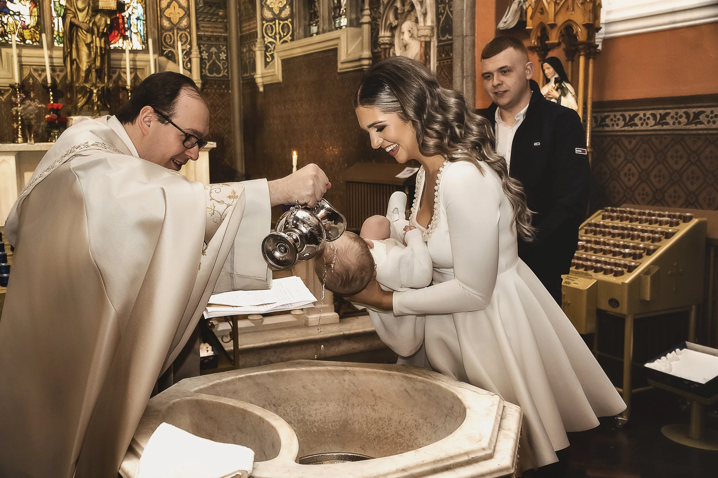 A religious baptism ceremony taking place in a church with intricate decorations. A priest in white robes is pouring water over a baby's head held by a woman in a white dress. Two young men are standing nearby, observing the ritual.