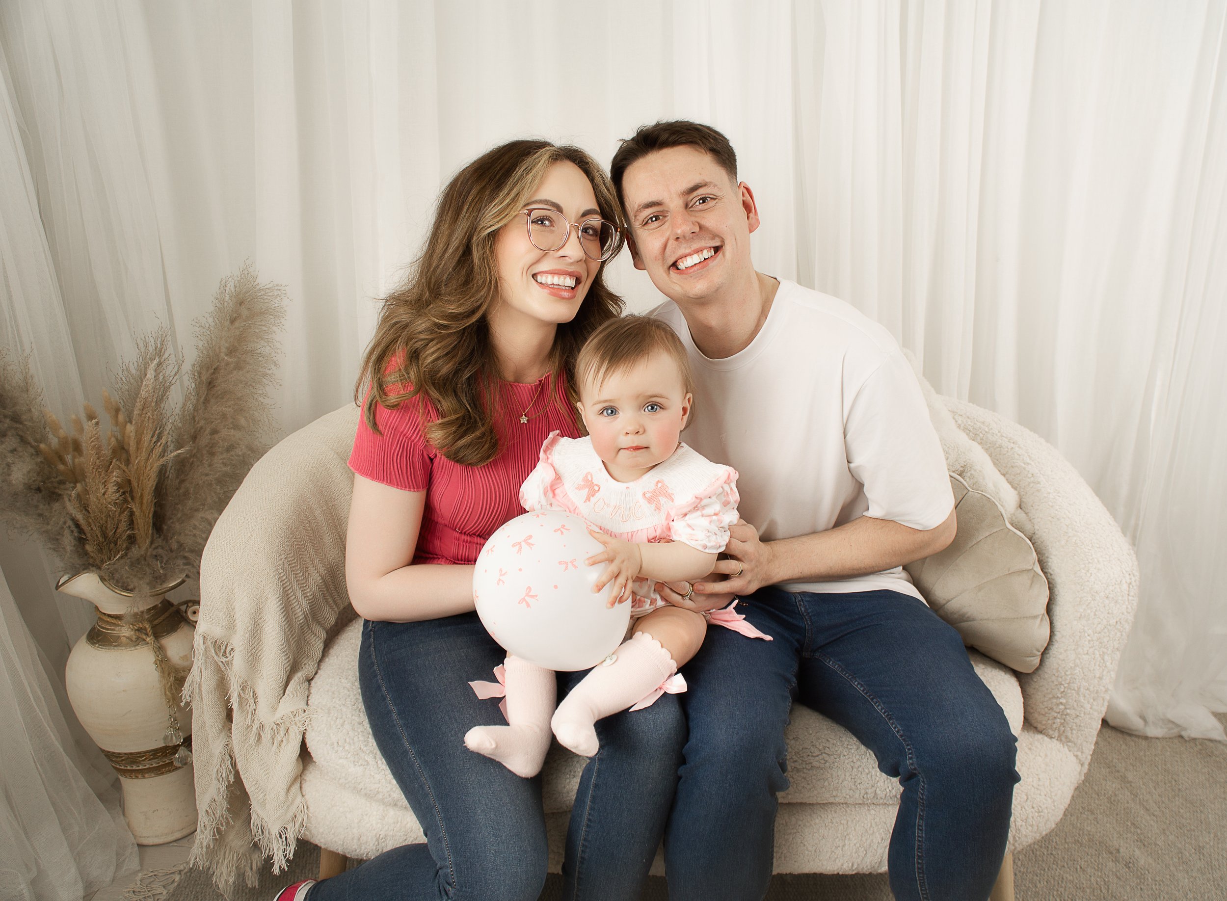 A smiling family of three, consisting of a woman with glasses, a man, and a baby girl, sitting on a beige sofa in a cozy room with white curtains and dried pampas grass.
