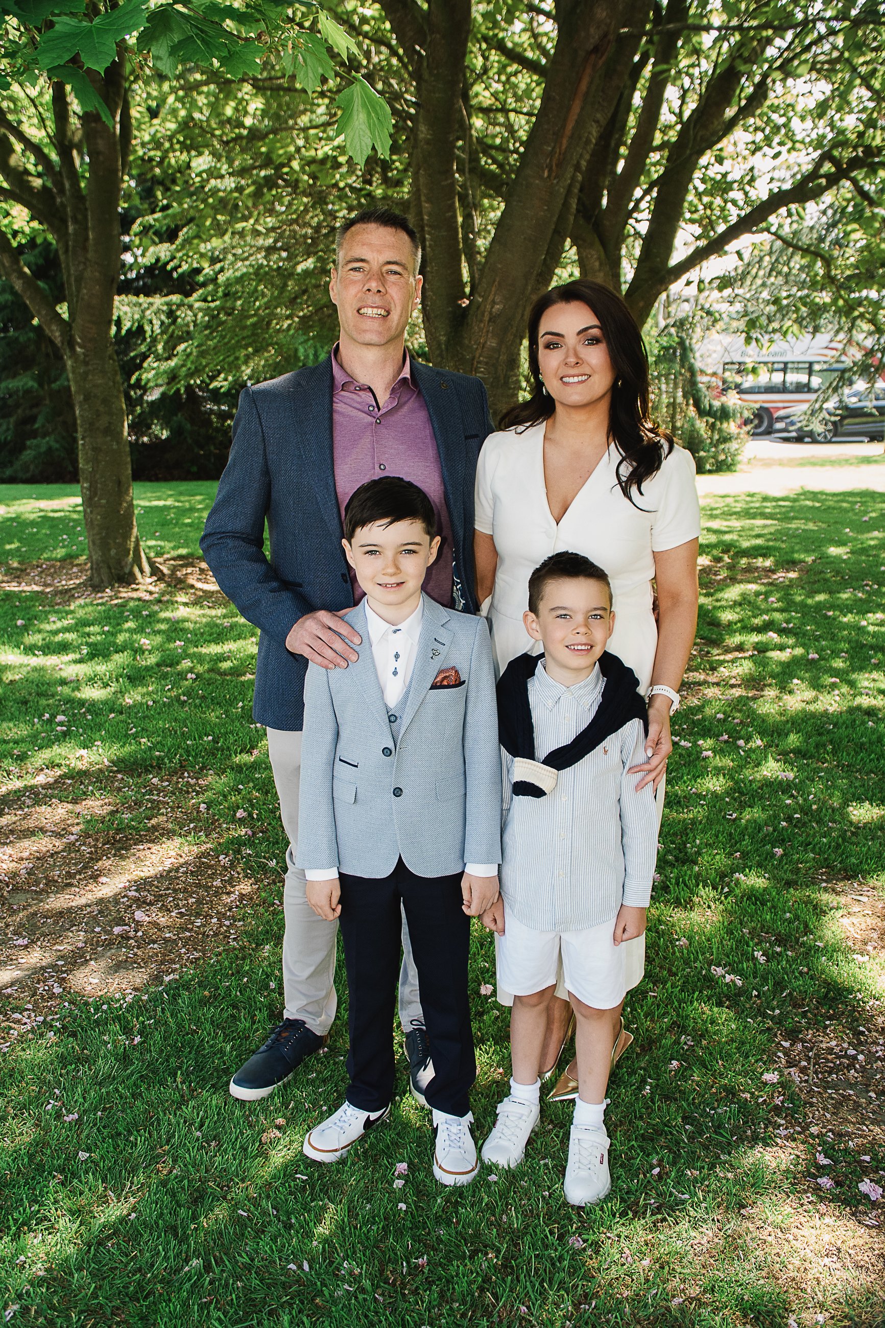 A family of five posing outdoors under a large leafy tree. Two adult males and females and two young boys, all dressed in semi-formal attire, standing on a grassy area with flower petals scattered around.