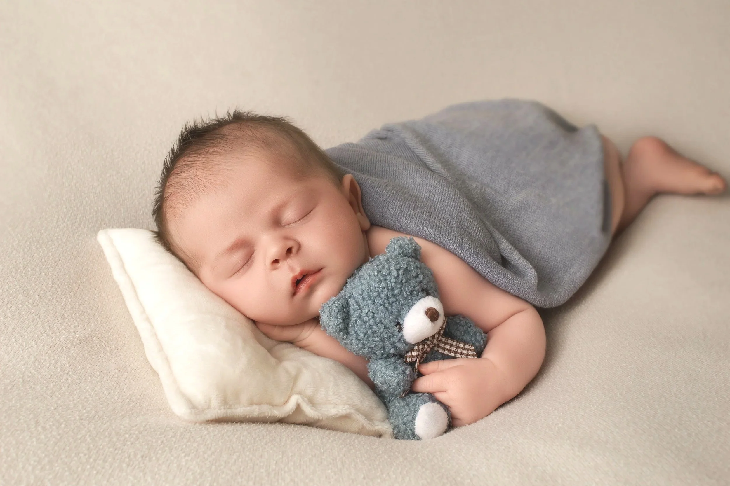 A sleeping baby lying on a soft beige surface with a cream pillow, holding a small plush gray teddy bear, wearing a gray shirt and patterned shorts.