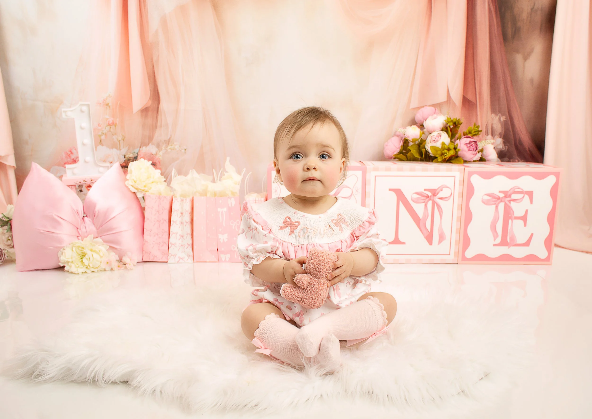 A young girl sitting on a fluffy white rug at a birthday celebration with pink and white decorations, holding a small pink stuffed bunny, with gift bags, flowers, and a large lit letter "L" in the background.