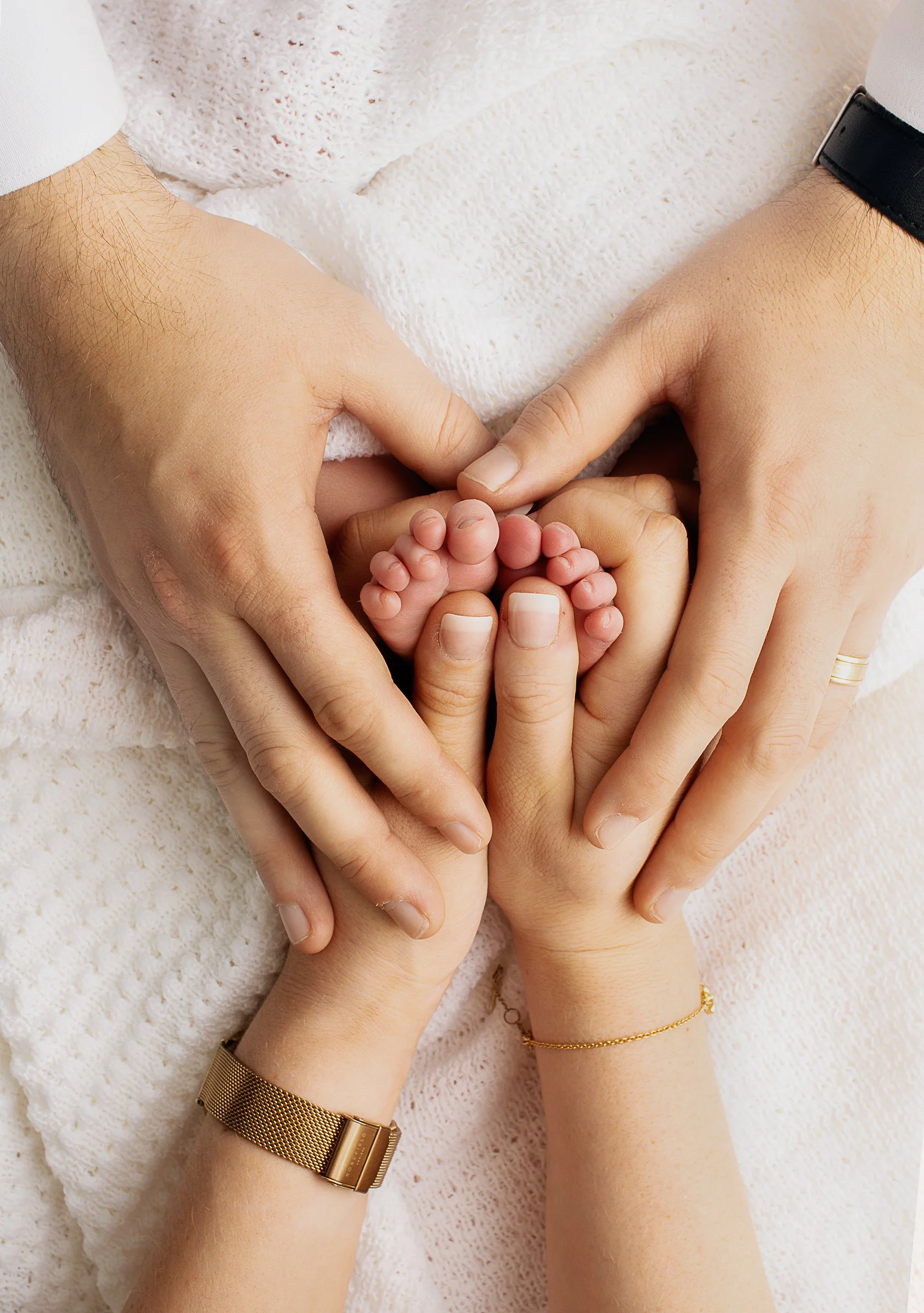 Two adults are holding a newborn baby's feet, creating a heart shape with their hands. The scene captures a tender moment of love and family bonding.