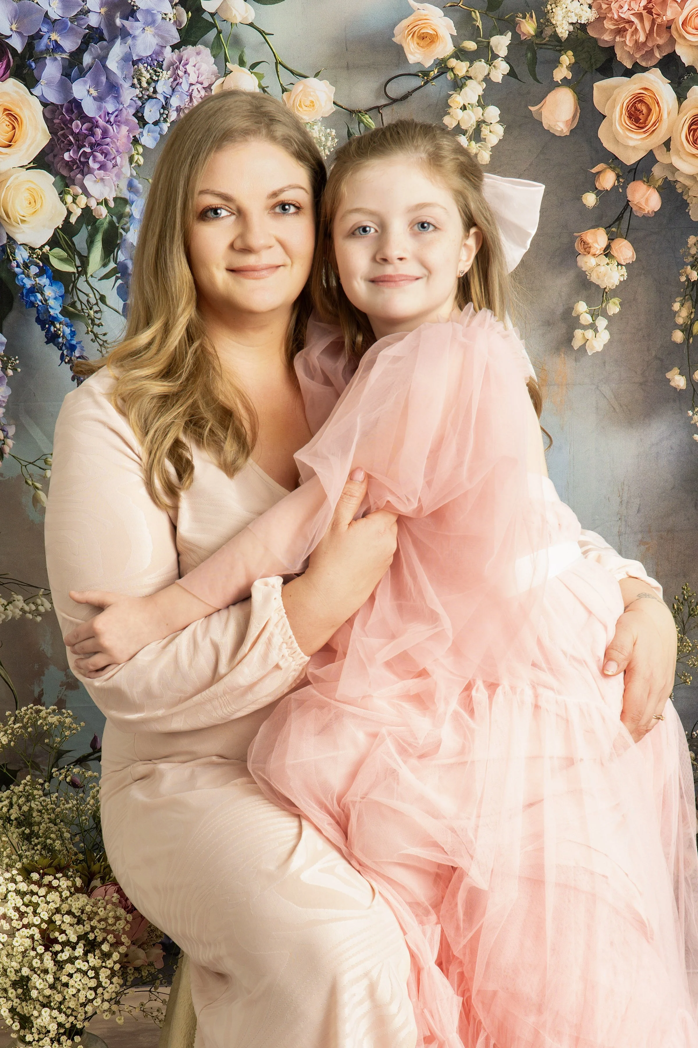 A woman and a young girl sitting close together, smiling, with a floral backdrop featuring roses, hydrangeas, and other flowers in pastel colors.
