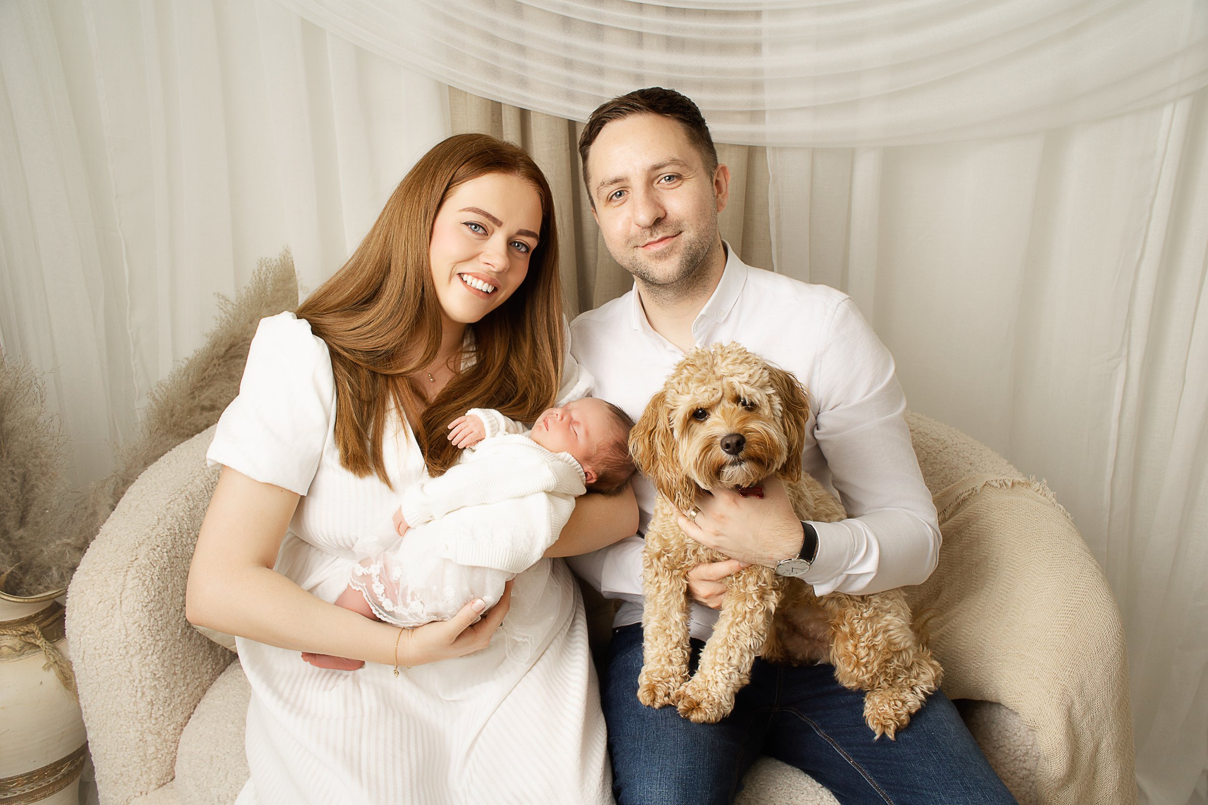A family of four with a dog and a newborn baby in a cozy, cream-colored living room.