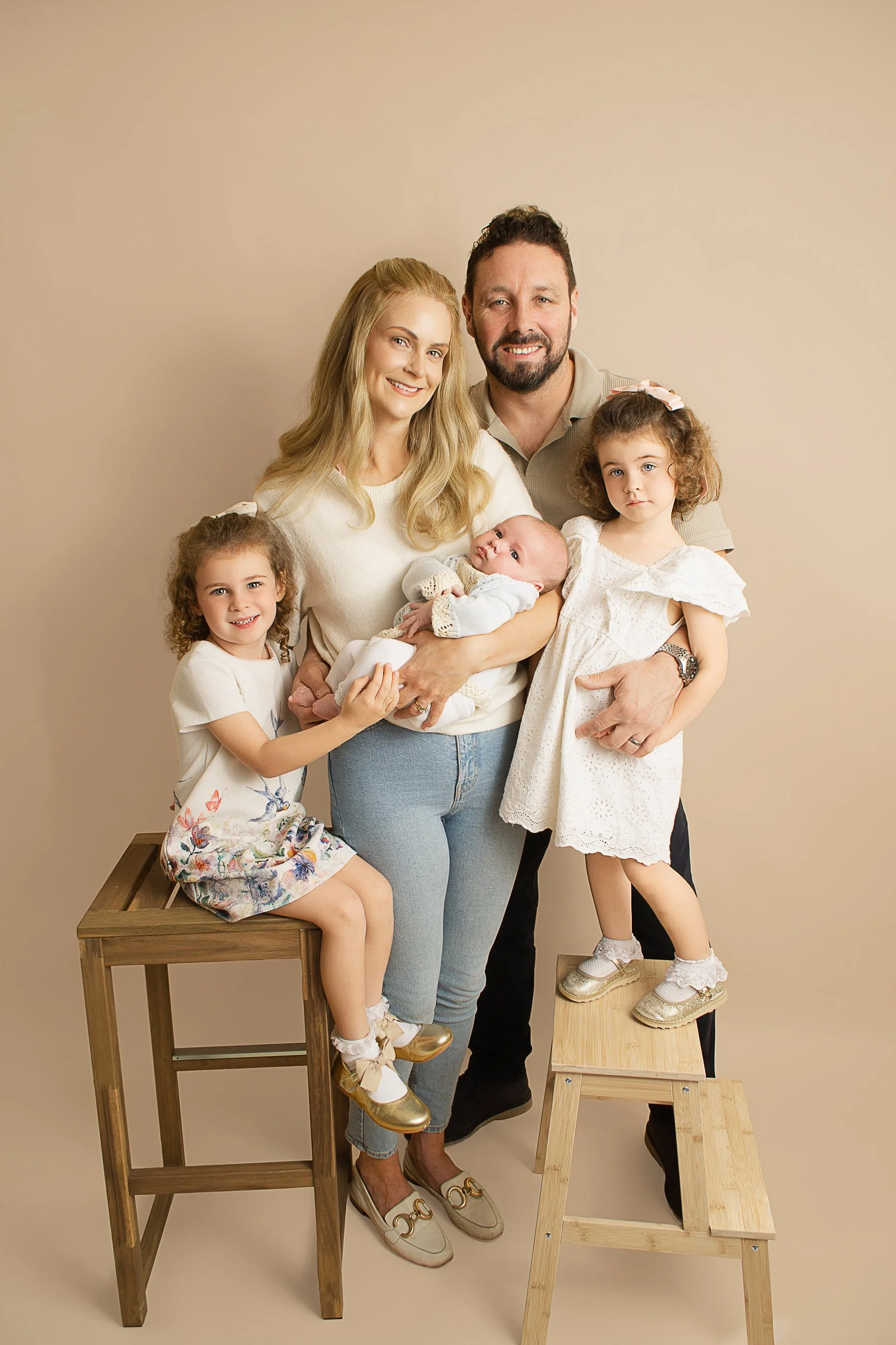 A family of five poses together indoors with a beige background. The mother is holding a baby, and there are two young girls standing on either side of her. They are all smiling, and the father is standing behind them.