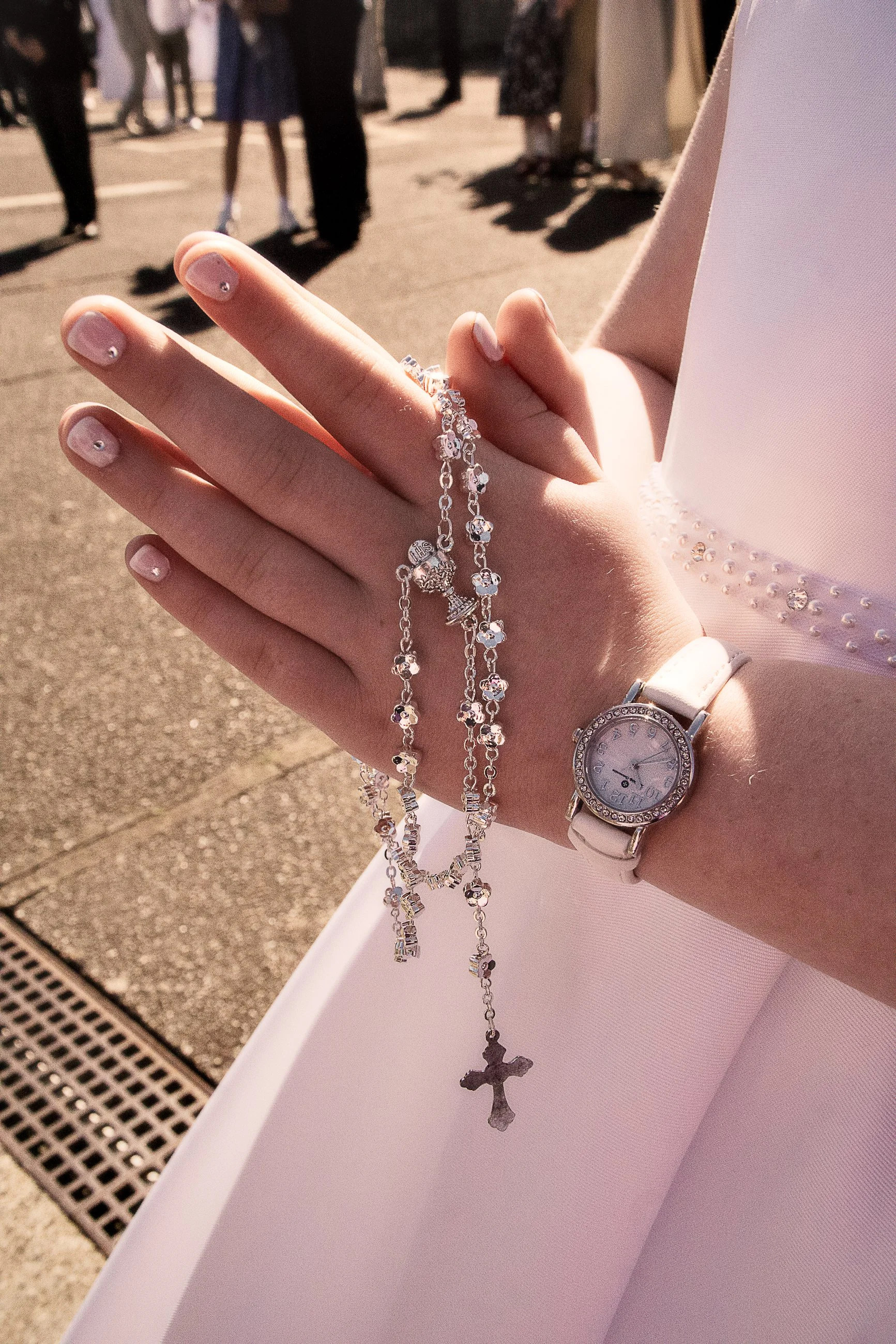 A close-up of a person's hand with a rosary featuring a cross pendant, jewelry, and a wristwatch. In the background, there are blurred people standing outdoors, likely at a gathering or event, on a sunny day.