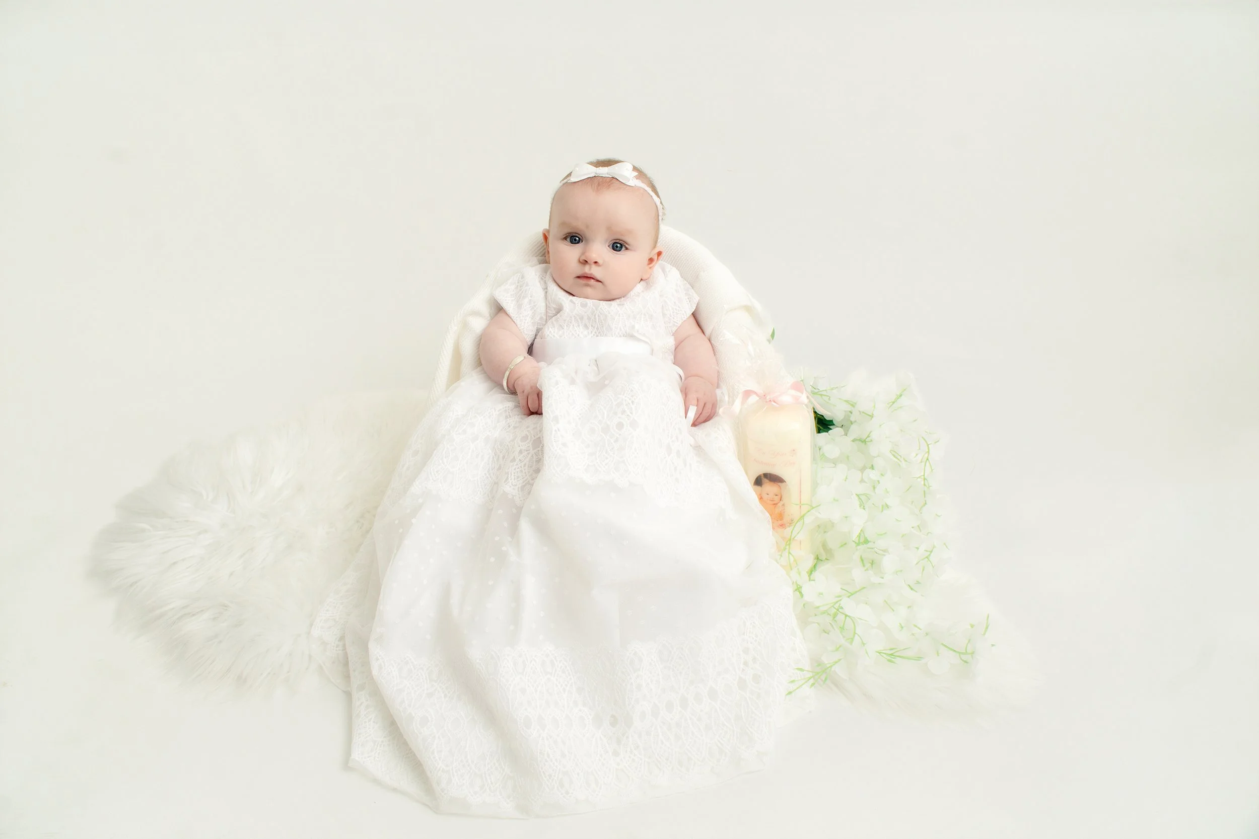 A baby girl dressed in a white lace dress sitting on a white background, with a birthday candle and flowers beside her.