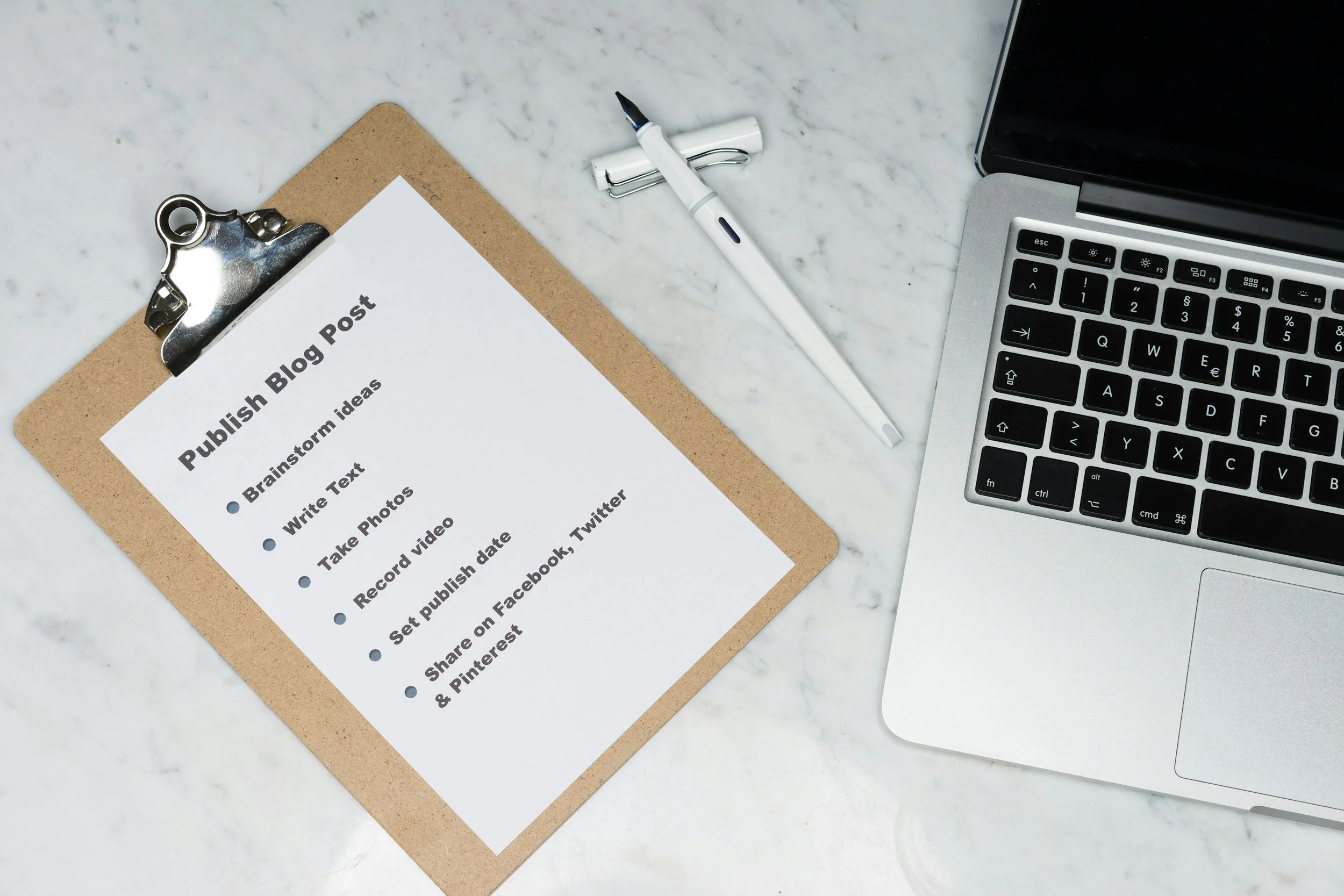A clipboard with a sheet of paper titled 'Publish Blog Post' on a marble desk. The paper lists steps like brainstorming ideas, writing text, taking photos, recording videos, and sharing on social media. Next to the clipboard lies a white marker, and to the right, part of a laptop keyboard and screen are visible.