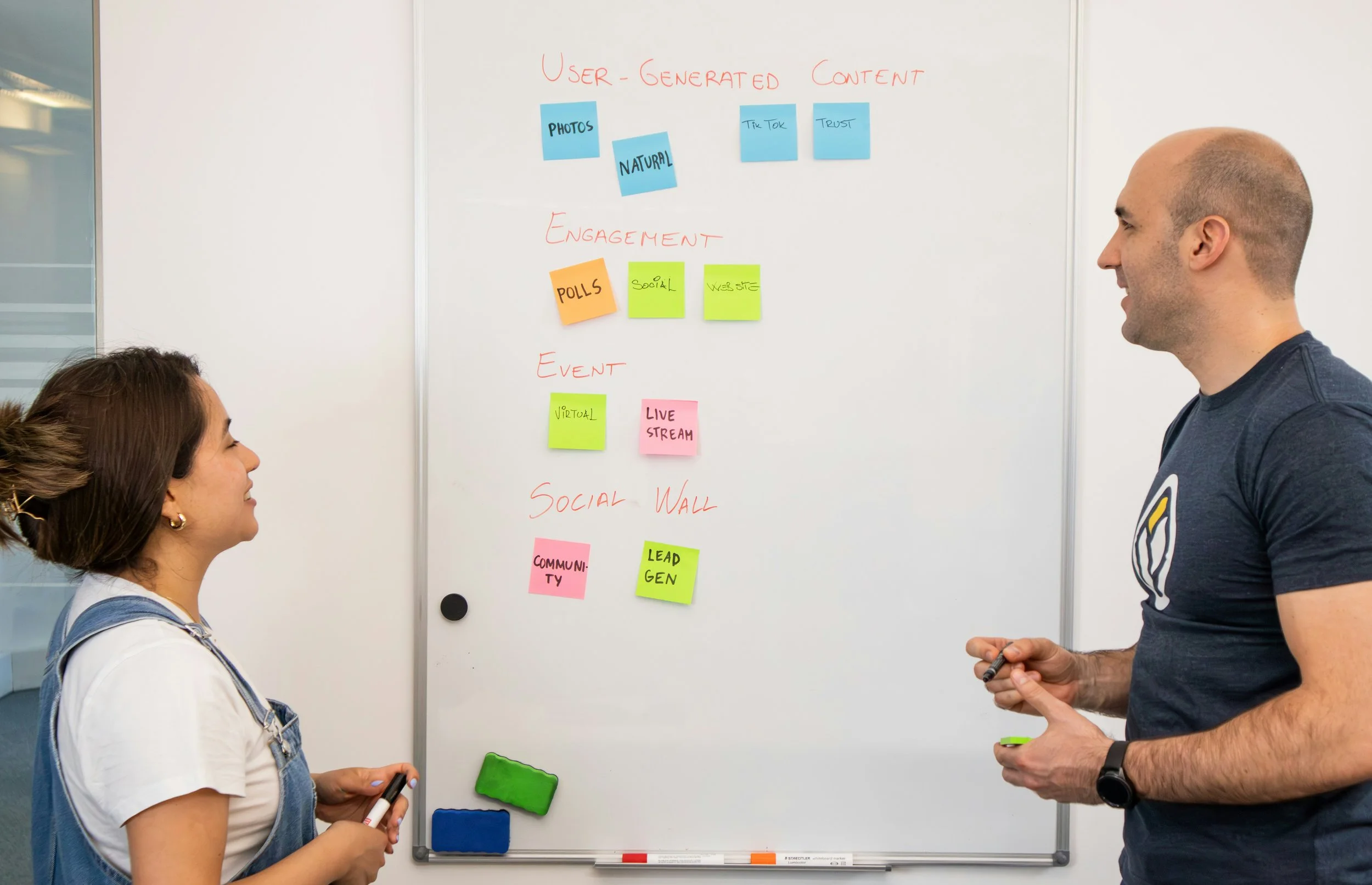 Two people standing in front of a whiteboard with notes about user-generated content, engagement, events, and social wall, with sticky notes on the board and one person holding a marker.