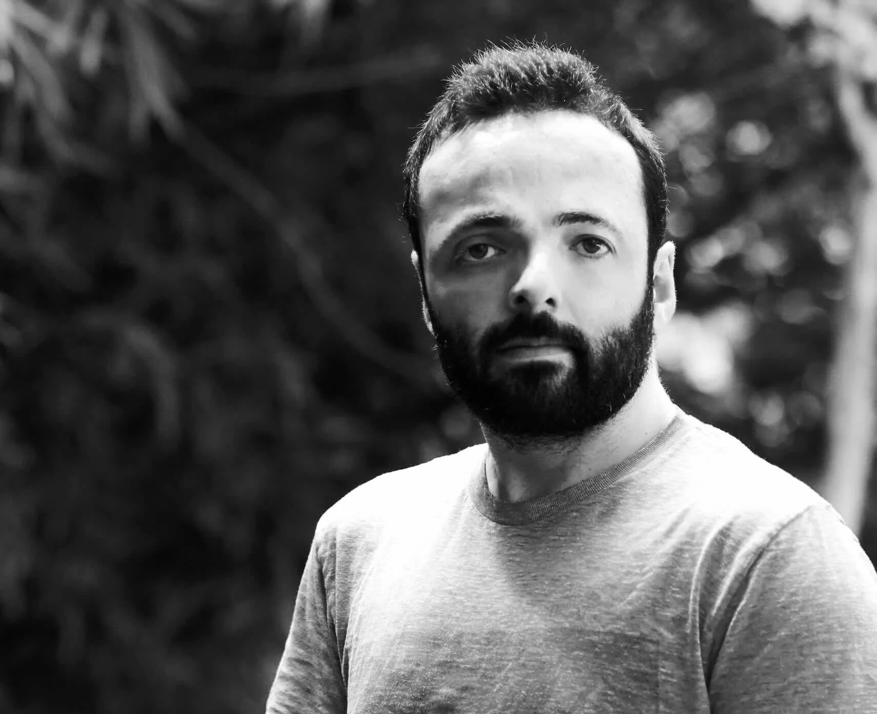 A black-and-white photo of a man with short curly hair, a full beard, and a mustache, wearing a light-colored t-shirt, standing outdoors with blurred foliage in the background.
