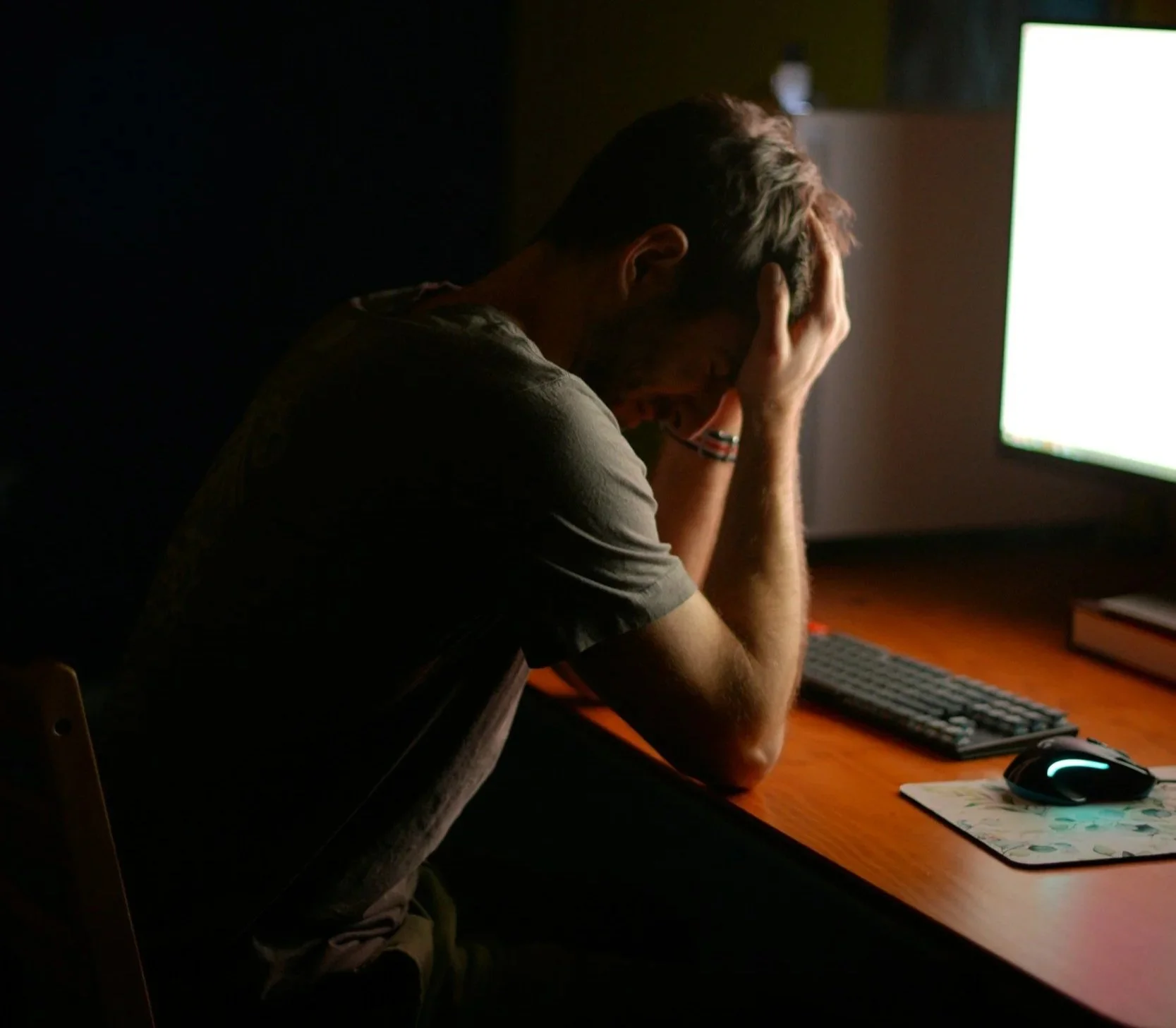 Person sitting at a desk in front of a computer, holding their head with one hand, appearing distressed or overwhelmed, with a gaming mouse, keyboard, and mat on the desk in a dimly lit room.