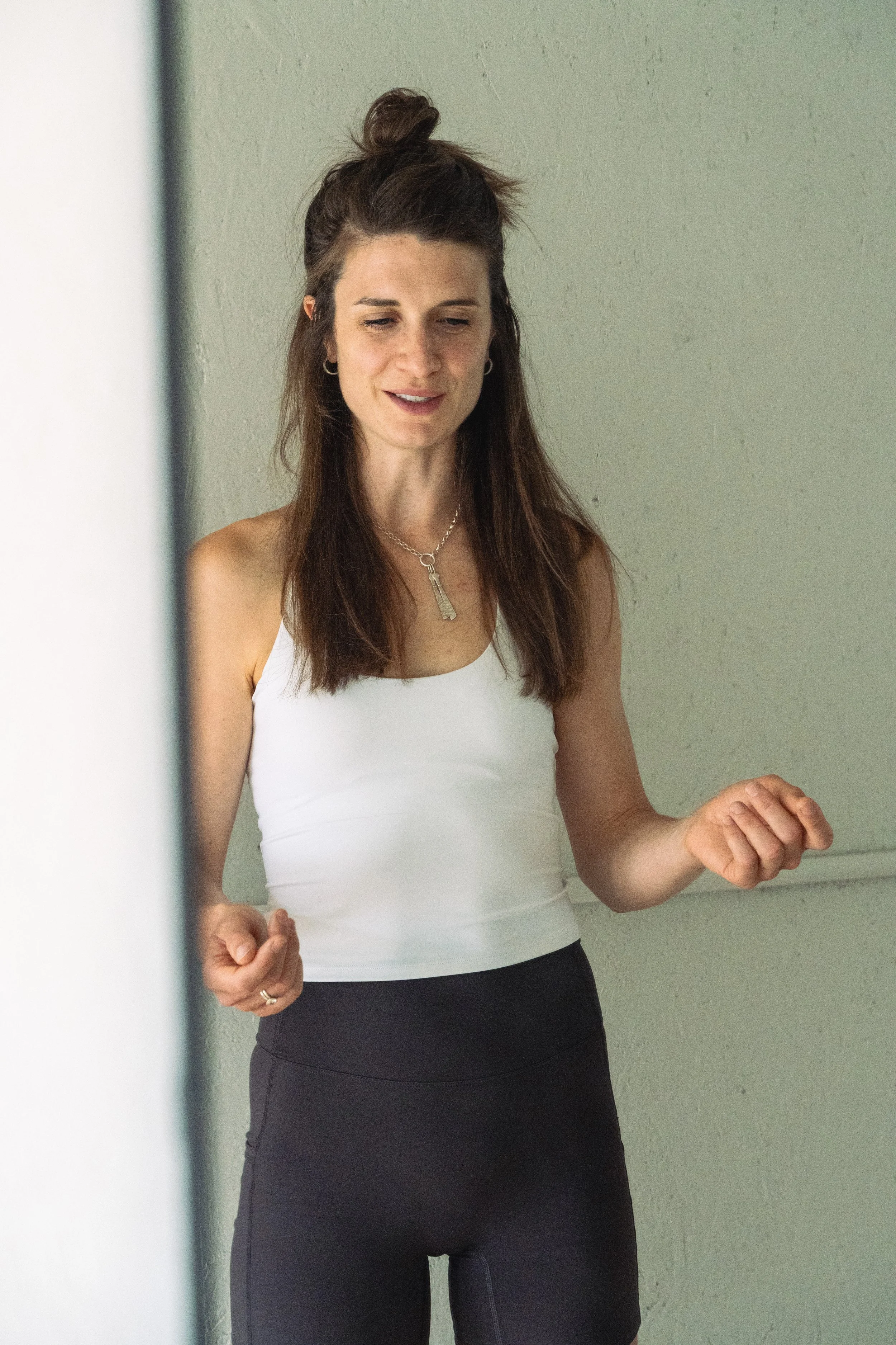 A woman with long brown hair in a half-up style, wearing a white sleeveless top and black athletic pants, appears to be exercising or stretching with clenched fists and slightly bent elbows, standing against a plain green wall.