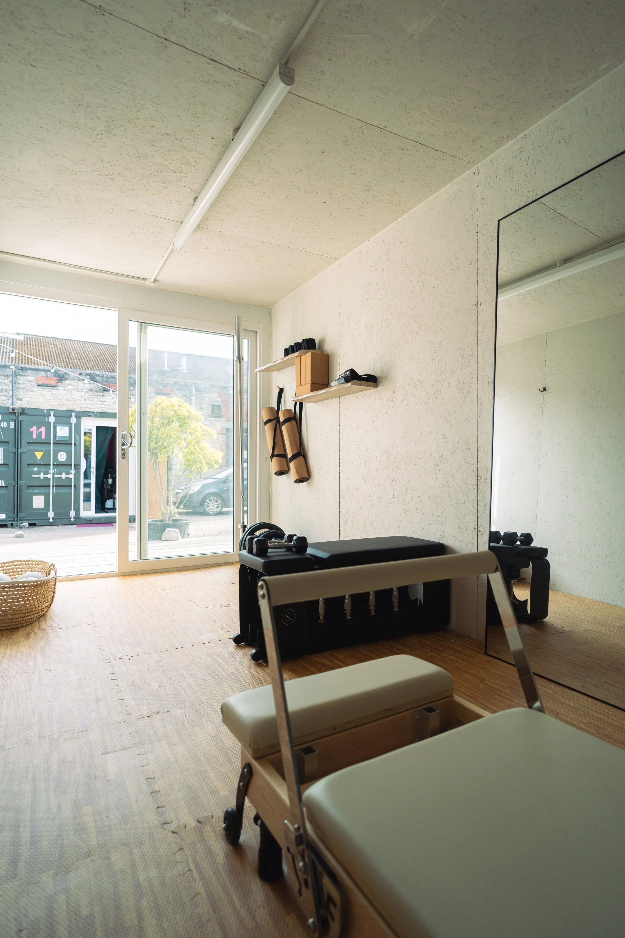 A medical or therapy room with a treatment table, a mirror on the wall, gym equipment, and shelves near a large window with a view outside of a green tree and buildings.