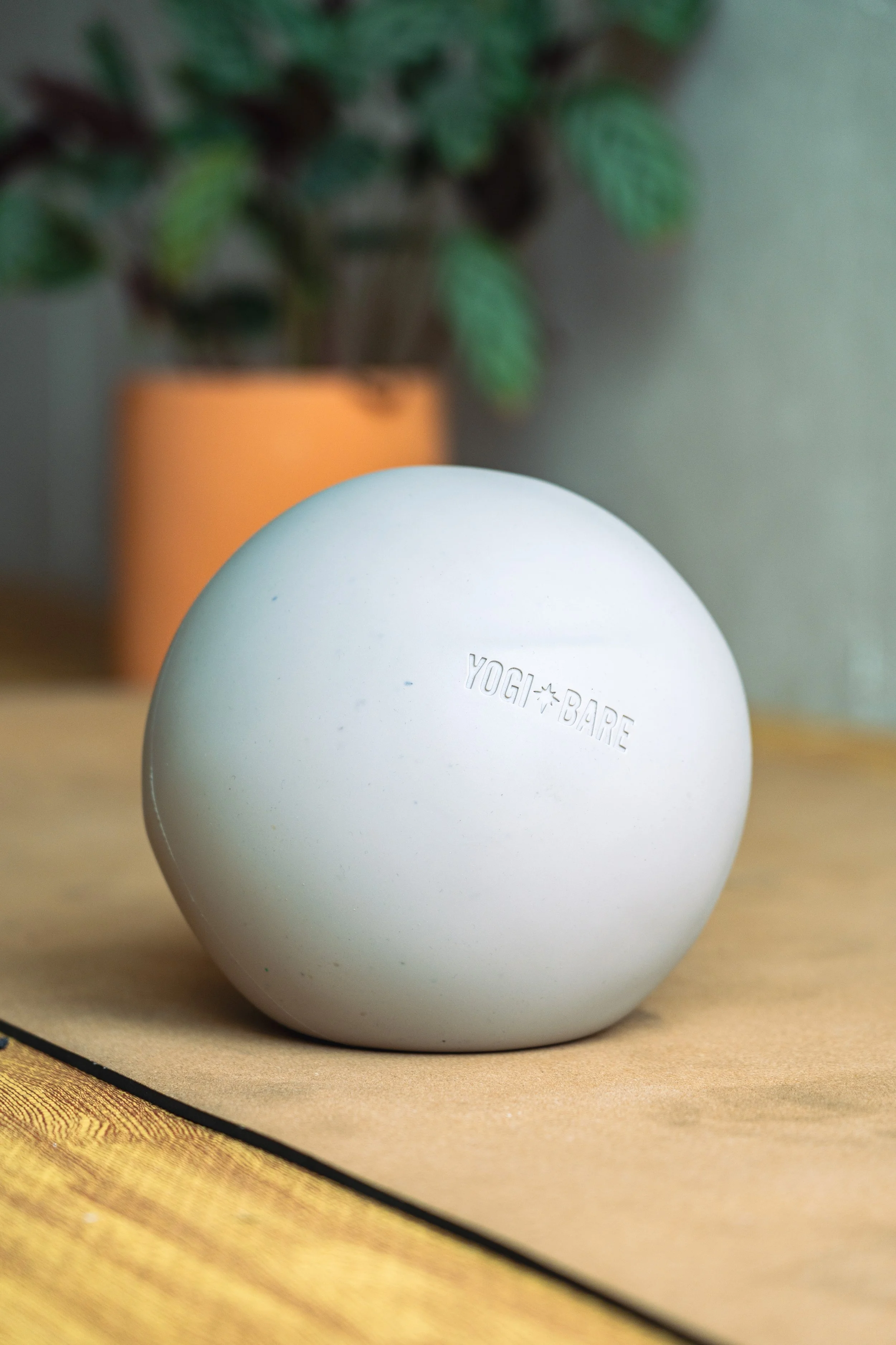Close-up of a white Yogi Bare egg-shaped skincare product in front of a potted plant on a wooden surface.