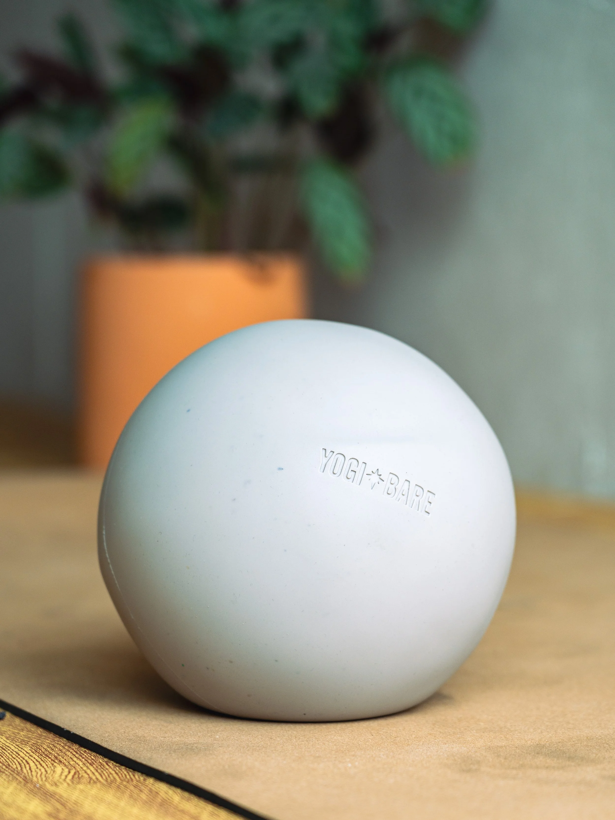 A white pilates ball with the words "YOGI★BARE" engraved on it, placed on a cork pilates mat on a wooden floor. In the background, there is a green leafy plant in an orange pot.