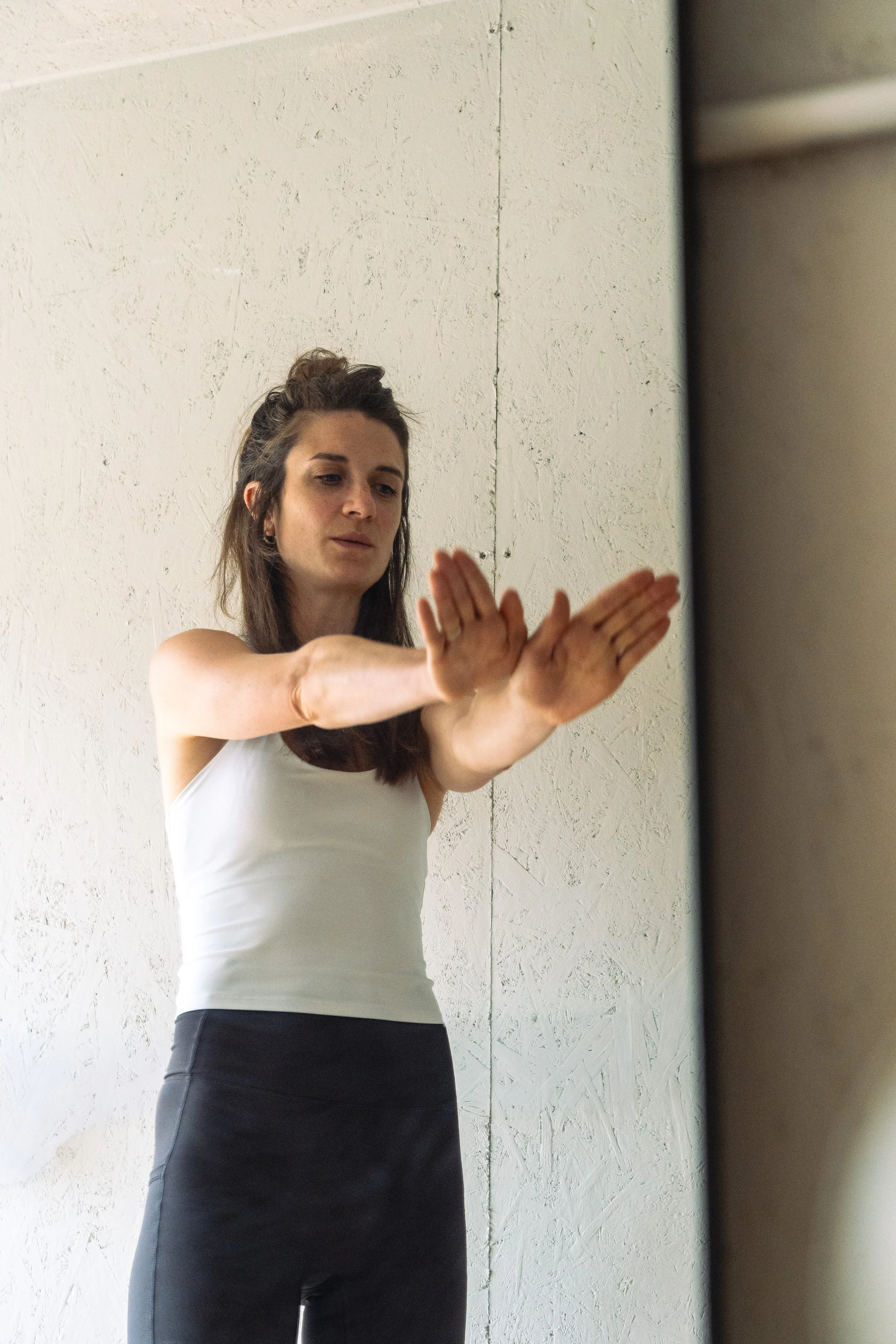 A woman practicing pilates or stretching in front of a mirror in a room with textured white walls, wearing a white tank top and black tights.