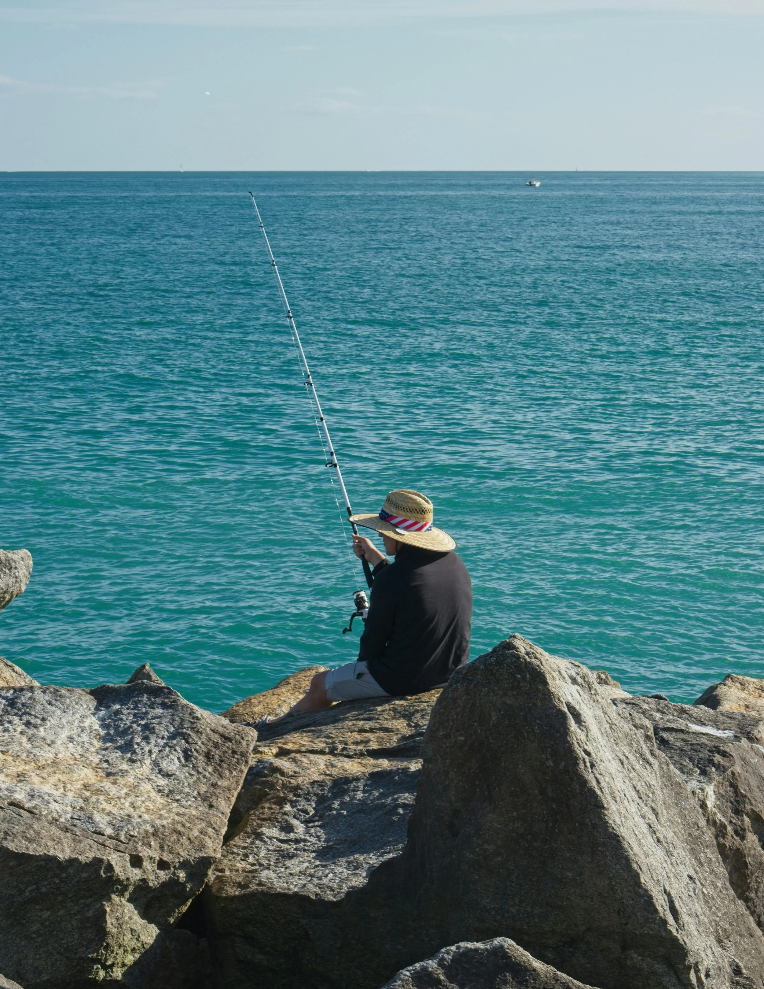 Person sitting on rocks near the water with a fishing pole, wearing a wide-brimmed hat, looking out at the ocean with boats in the distance.