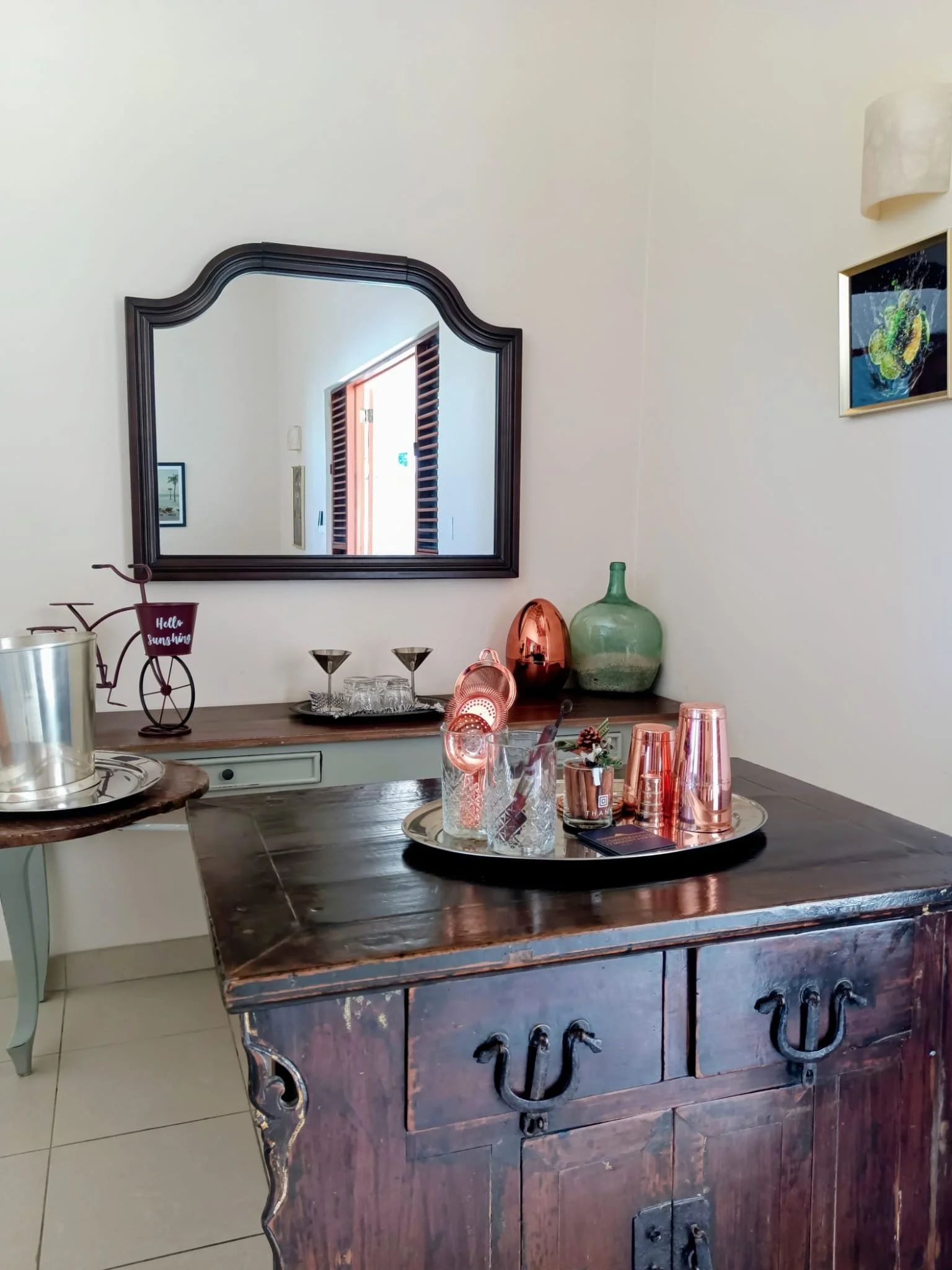 Interior view of a wooden sideboard with decorative metal handles, topped with glassware, copper cups, and a plate with red cups. A large mirror with a dark frame hangs above the sideboard, reflecting a doorway with wooden shutters and a picture on the wall.