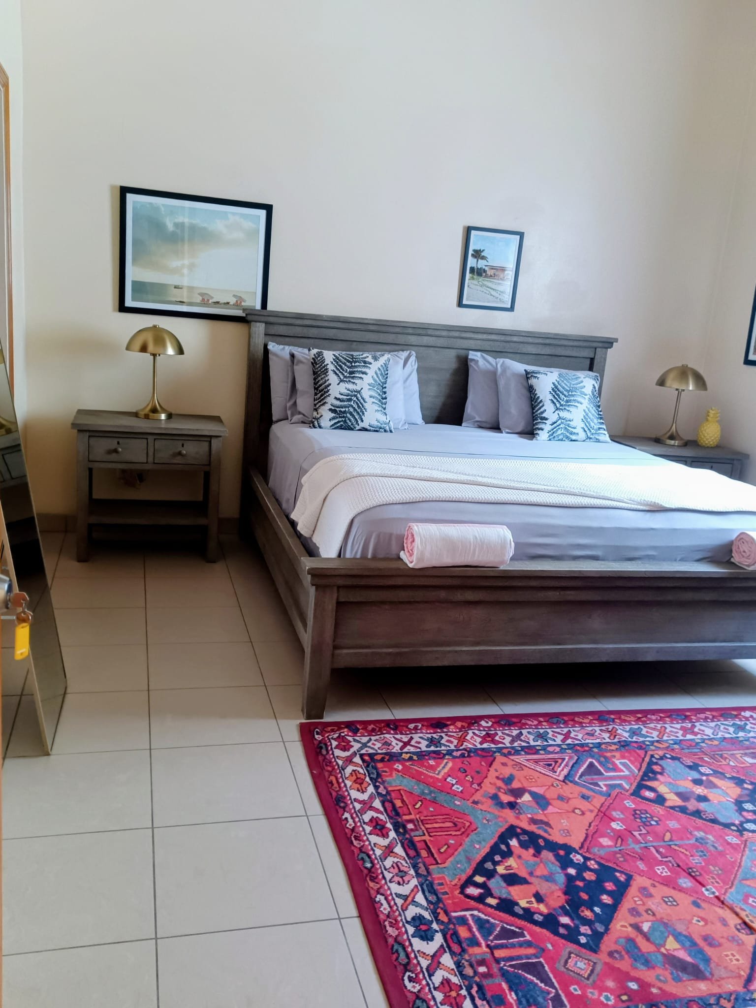 A bedroom with a bed, two wooden nightstands with lamps, two framed pictures on the wall, a red patterned rug, and a rolled towel at the end of the bed.