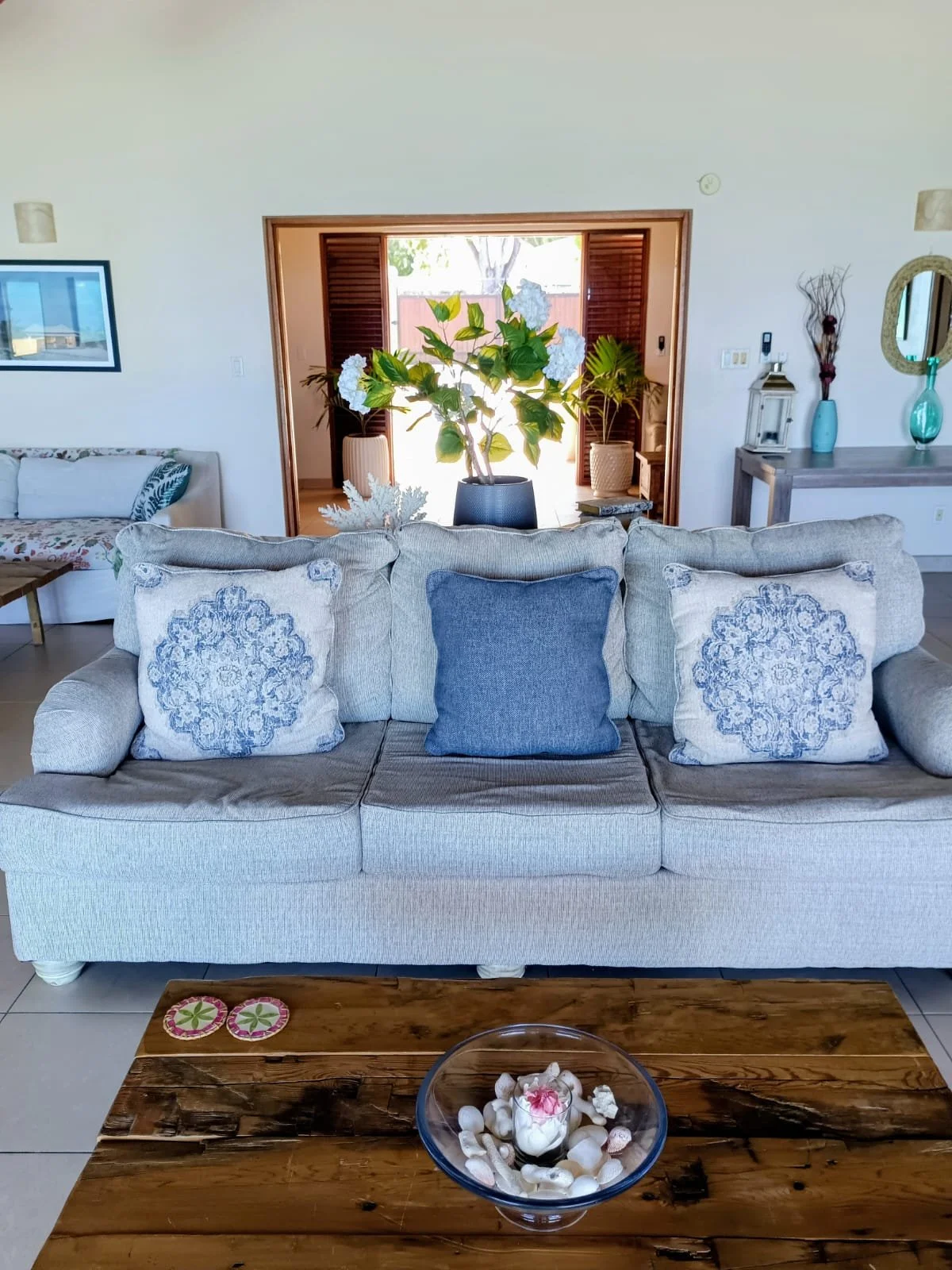 Living room with beige sofa, blue and patterned pillows, wooden coffee table with shell decor, potted plant in the background near open door, other furniture and decorative items visible.