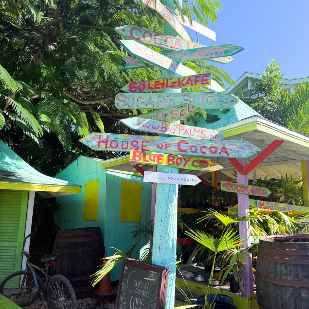 Colorful wooden directional signs pointing to various shops and locations, surrounded by tropical plants, beneath a bright blue sky. The signs include names like Cocoa, Söle Kafe, Sugar & Spice, House of Cocoa, Blue Boy, and others.