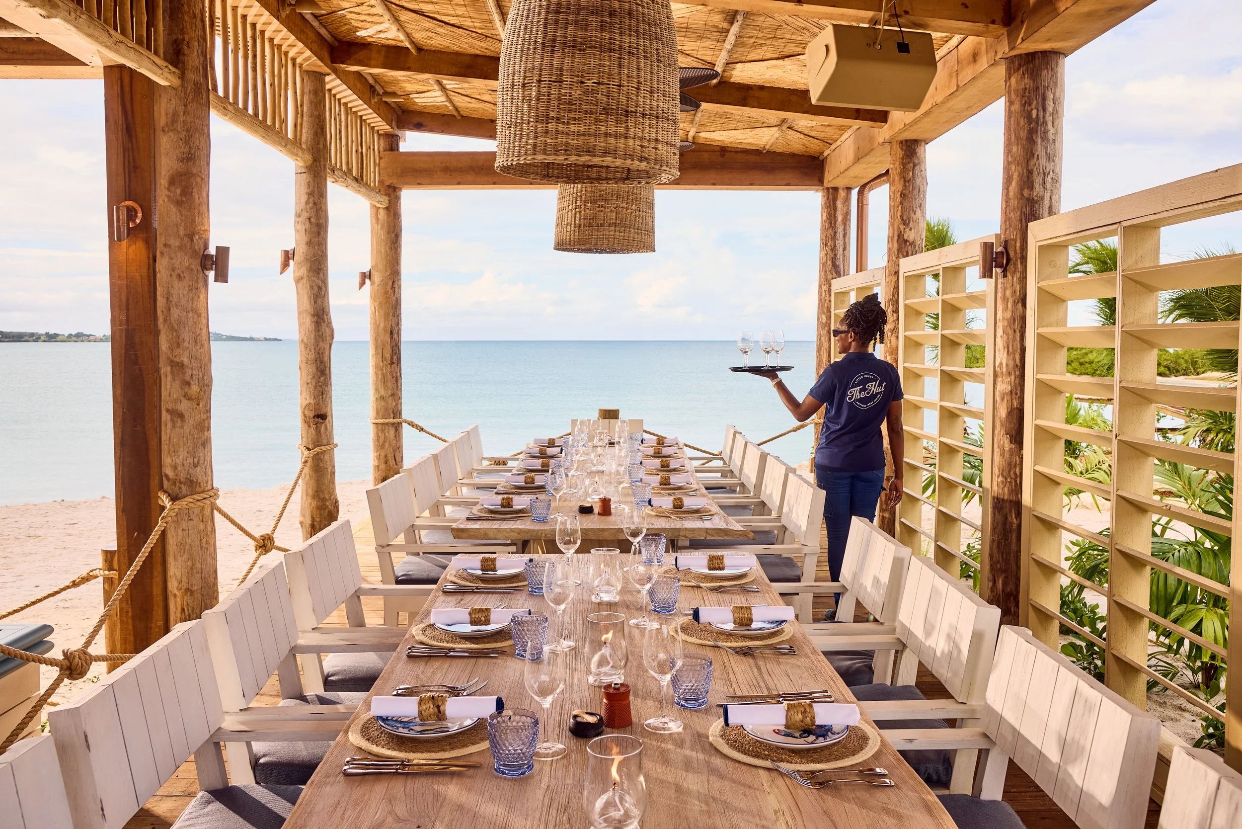 Long wooden table set with wine glasses, water glasses, plates, napkins, and silverware, located outdoors on a beach under a wooden canopy, with a view of the ocean and a cloudy sky in the background. A server carrying a tray of wine glasses is walking beside the table.