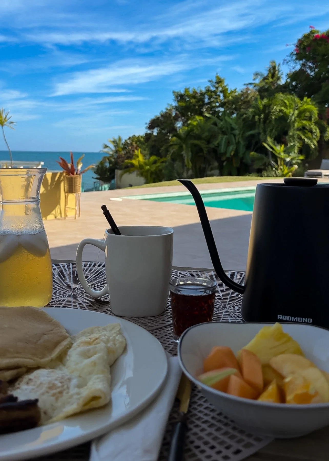 Breakfast setup outdoors with a view of a swimming pool, ocean, and greenery, including a pitcher of lemonade, a mug, a small jar of jam, a plate of eggs and flatbread, and a bowl of cut fruits.