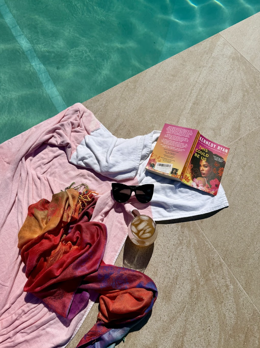 Poolside scene with a pink and white towel, colorful scarves, black sunglasses, a glass of iced coffee, and a book titled 'This Could Be Us' by Kennedy Ryan.