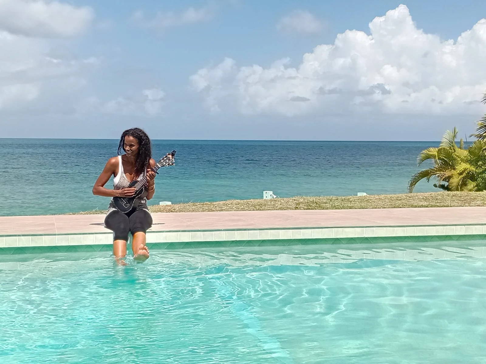 Woman sitting on pool edge playing ukulele with ocean and clouds in background.