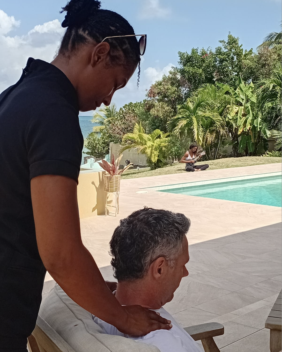 A man giving a massage to another man sitting on a lounge chair beside a swimming pool, with tropical plants and a woman playing guitar in the background.