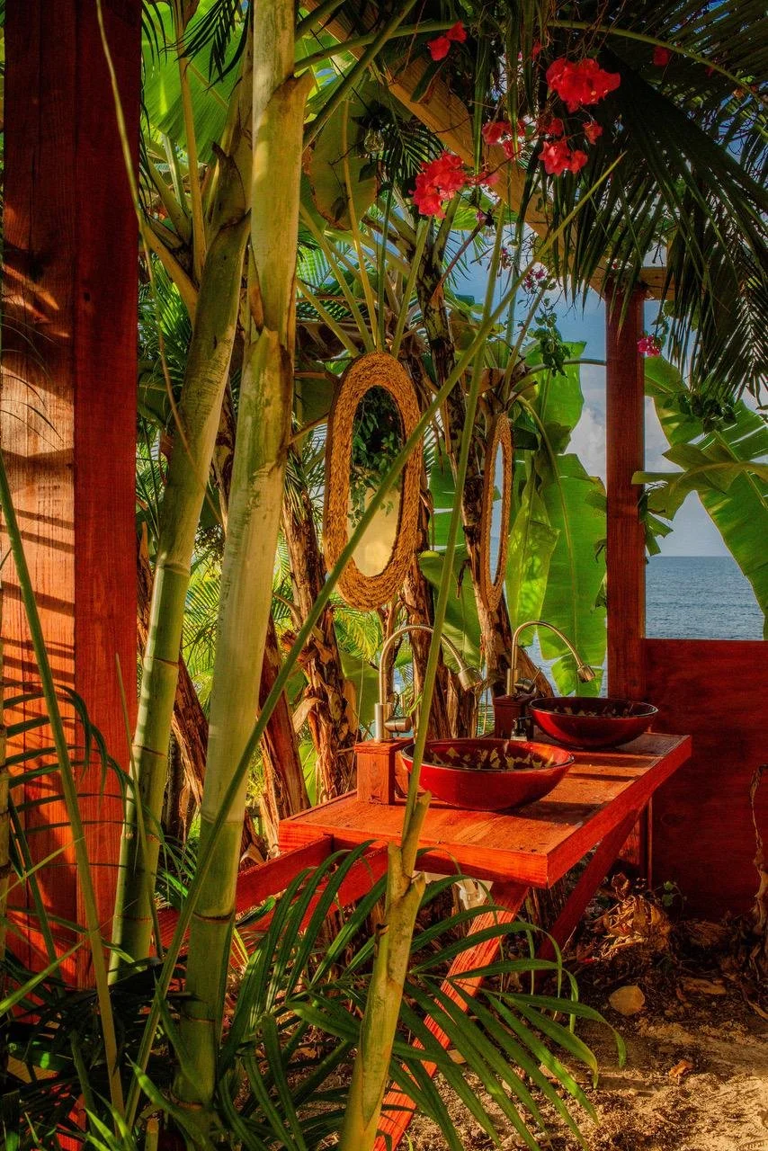 Tropical outdoor sink area with two black vessel sinks, wooden countertop, surrounded by lush green foliage, pink flowers, and ocean view in the background.