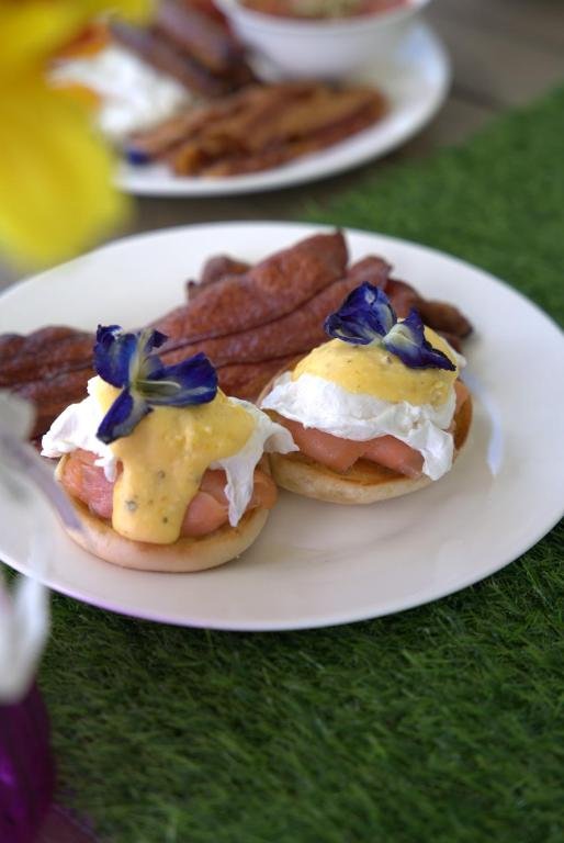Eggs Benedict topped with blue edible flowers, served with bacon on a white plate.