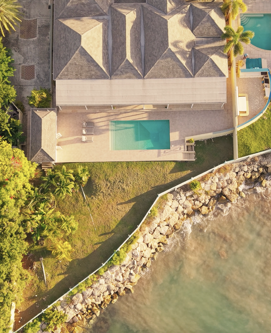 An aerial view of a house with a backyard that includes a swimming pool, lounging chairs, and surrounding lush greenery, bordered by rocks and water.