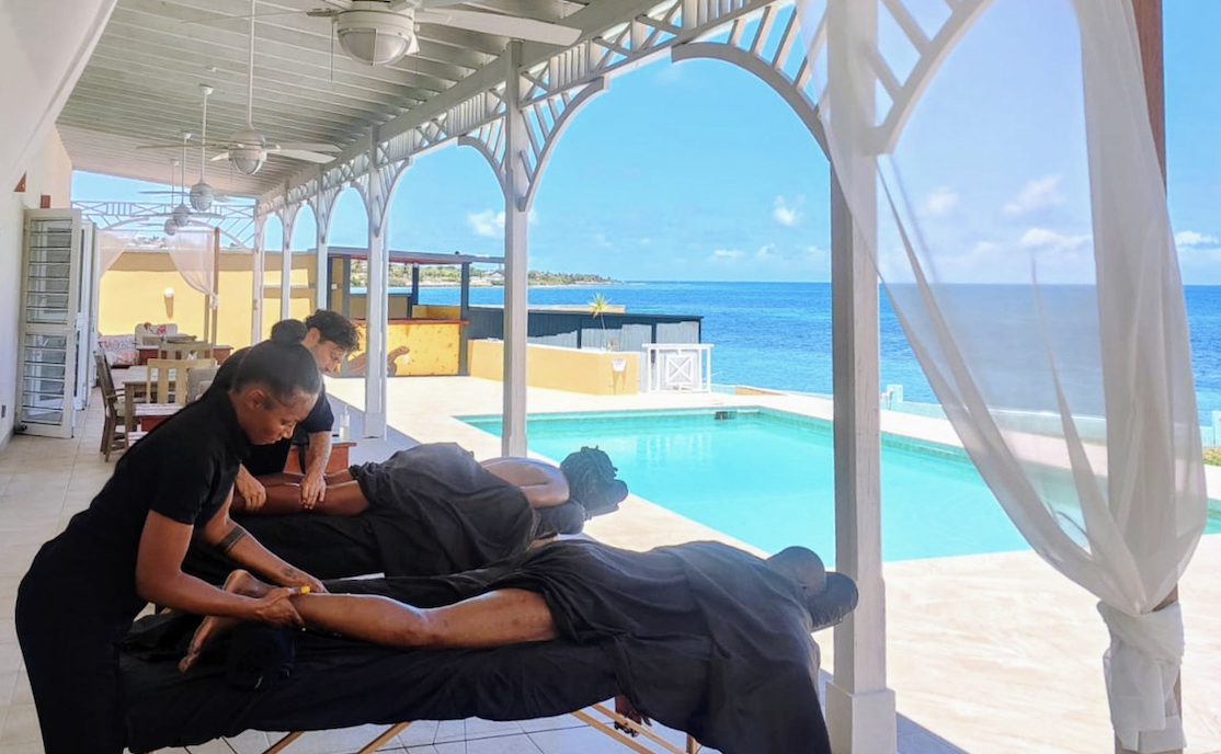 Massage therapists giving massages on outdoor massage tables by a pool, with ocean view and blue sky in the background.